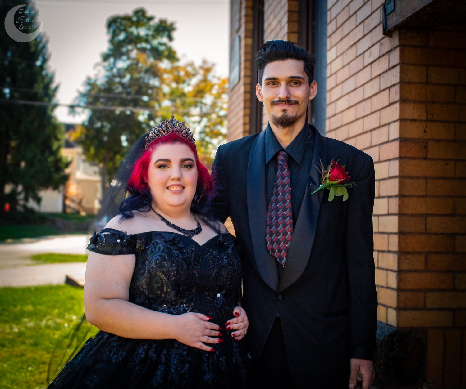 A couple in formal attire poses together outdoors against a brick building with autumn foliage in the background.