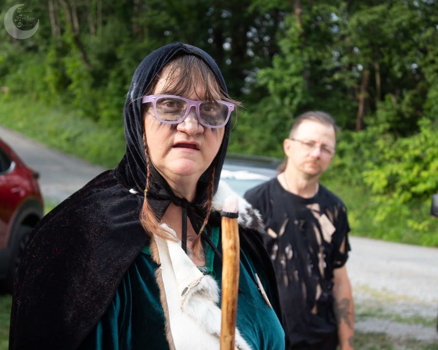 Two people stand on a rural road, one wearing a black cape and glasses in the foreground with trees in the background.
