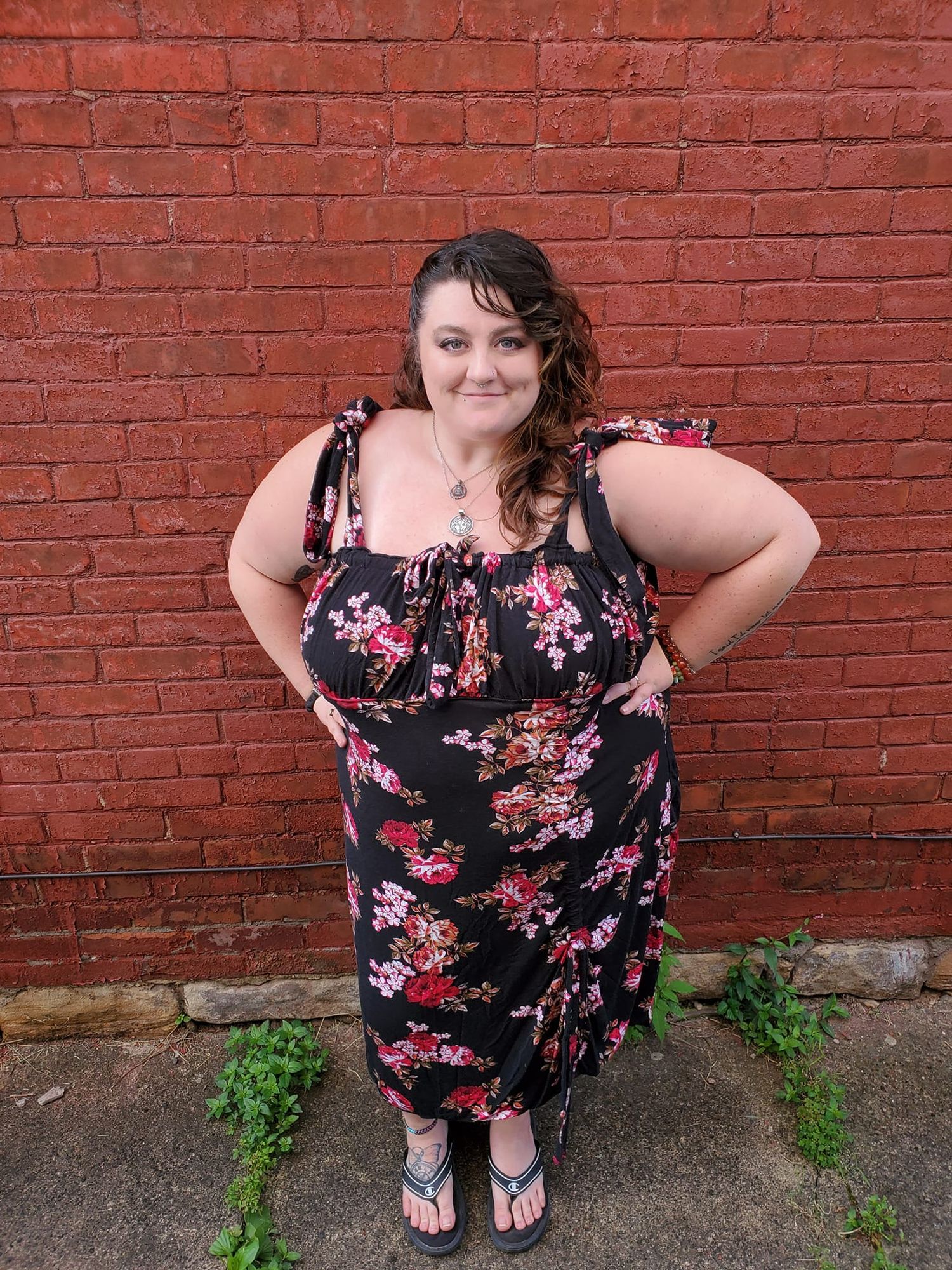 A person in a floral dress poses confidently against a red brick wall background.