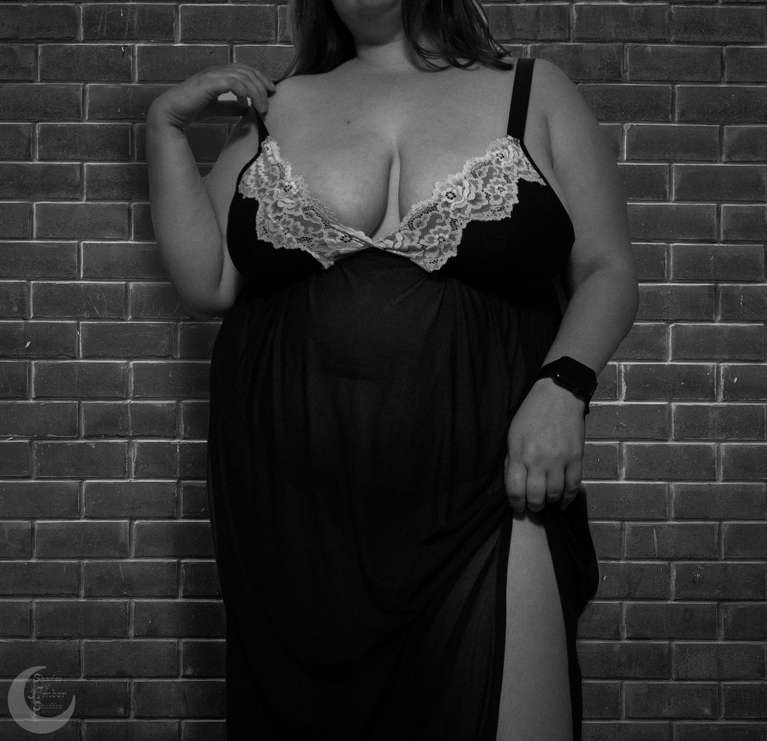 Curvy figure in black negligee with white lace trim poses against brick wall in black and white photograph.