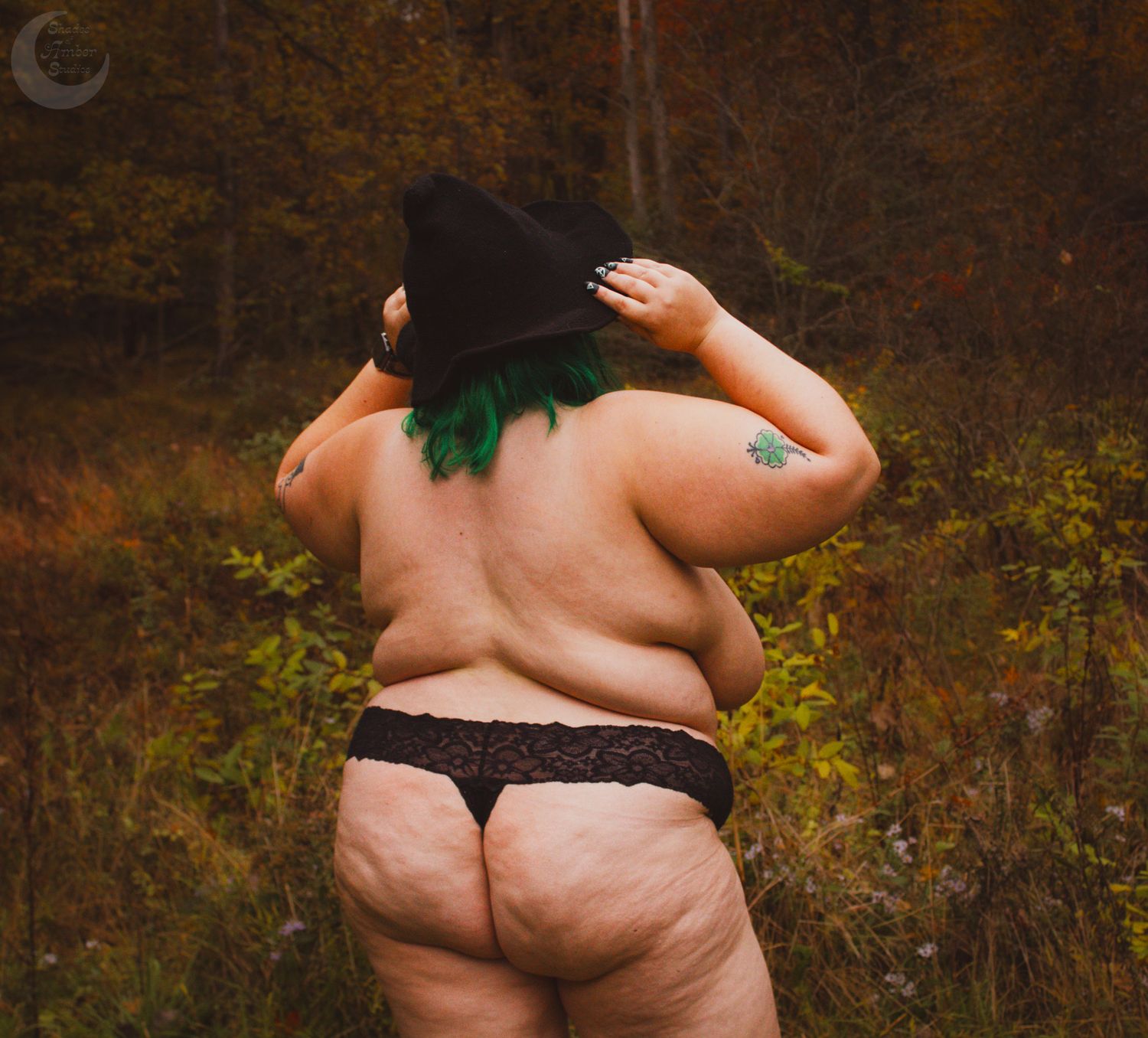 Plus size figure in black lingerie and witch hat poses in autumn forest setting.