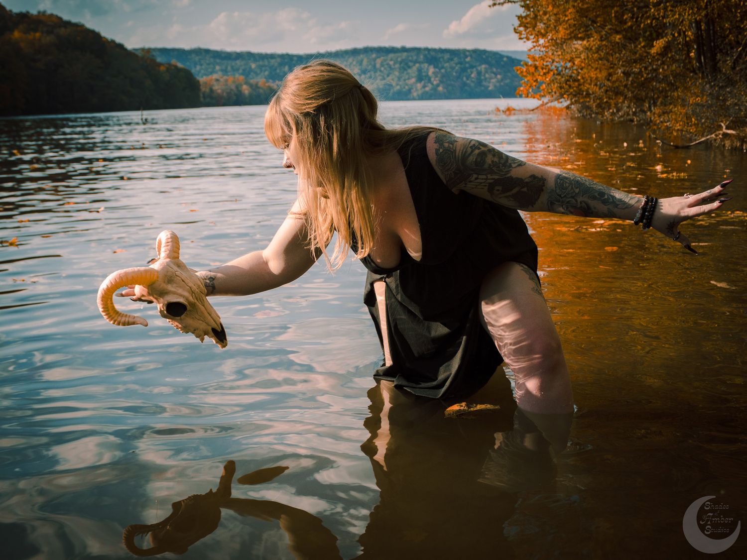 Woman in dark clothing reaches into a lake at sunset with golden light reflecting off the water.