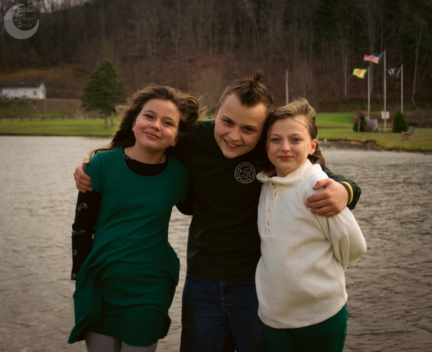 Three people stand together smiling by a lake on an overcast day, wearing casual sweaters.