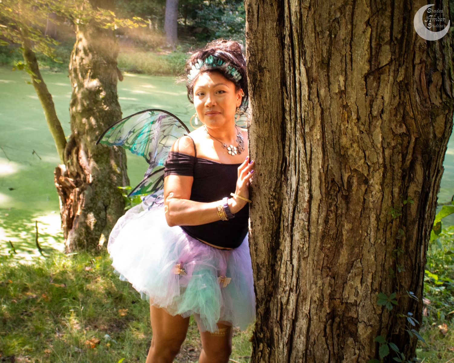 A figure in a tulle fairy costume leans against a large tree trunk near a green pond in a dreamy setting.