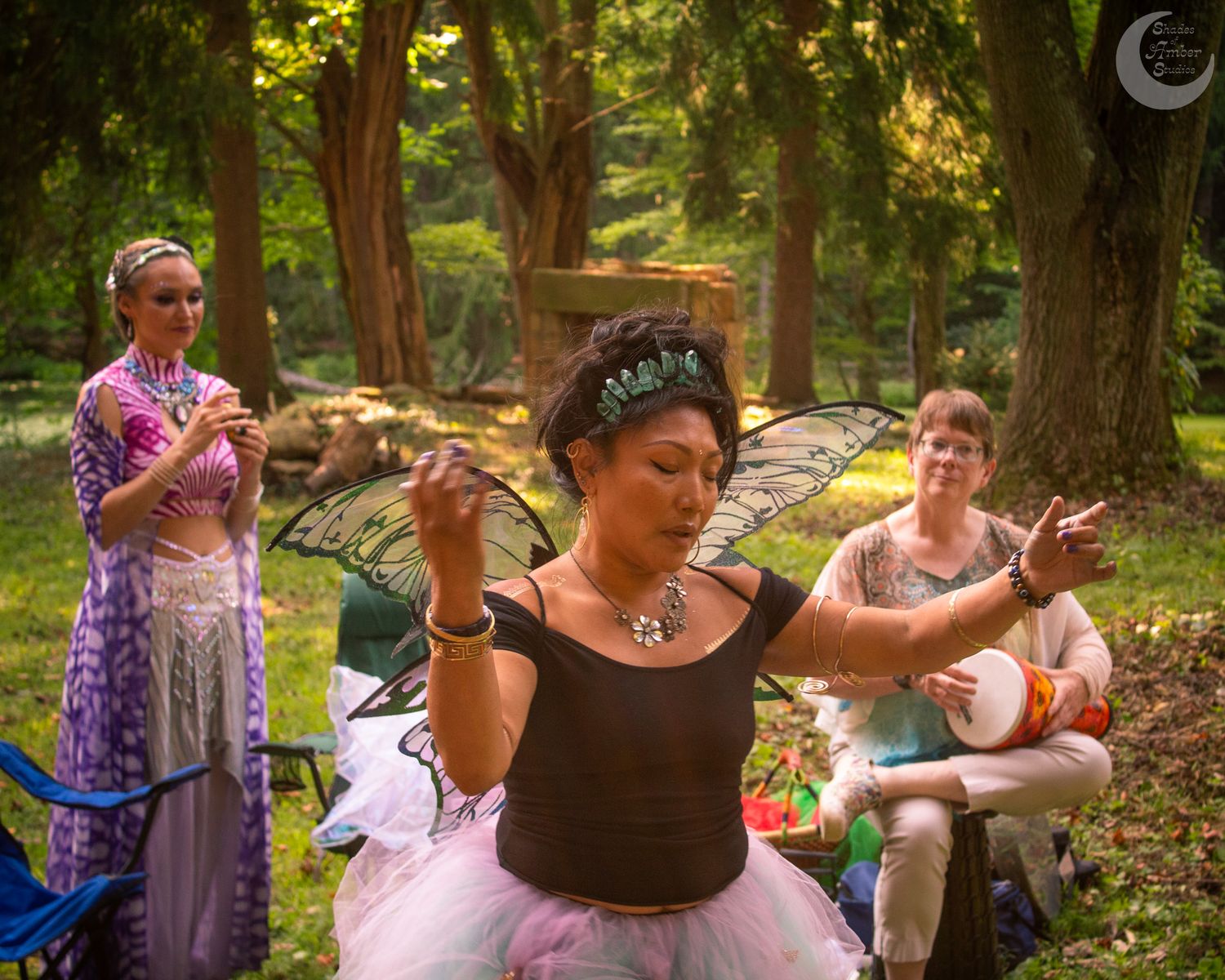 A group wearing rustic clothing and steaming cups gathers outdoors under a forest canopy during a woodland tea ceremony.