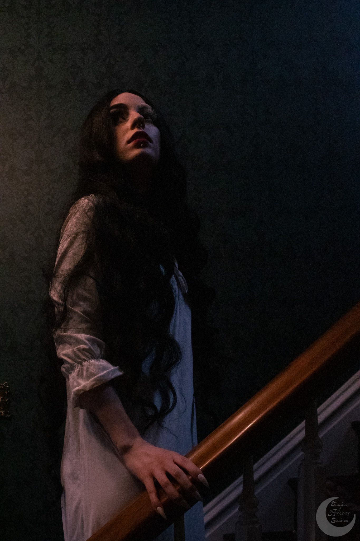 A pale woman in a white dress with long dark hair stands on a dimly lit staircase in a haunted gothic setting.
