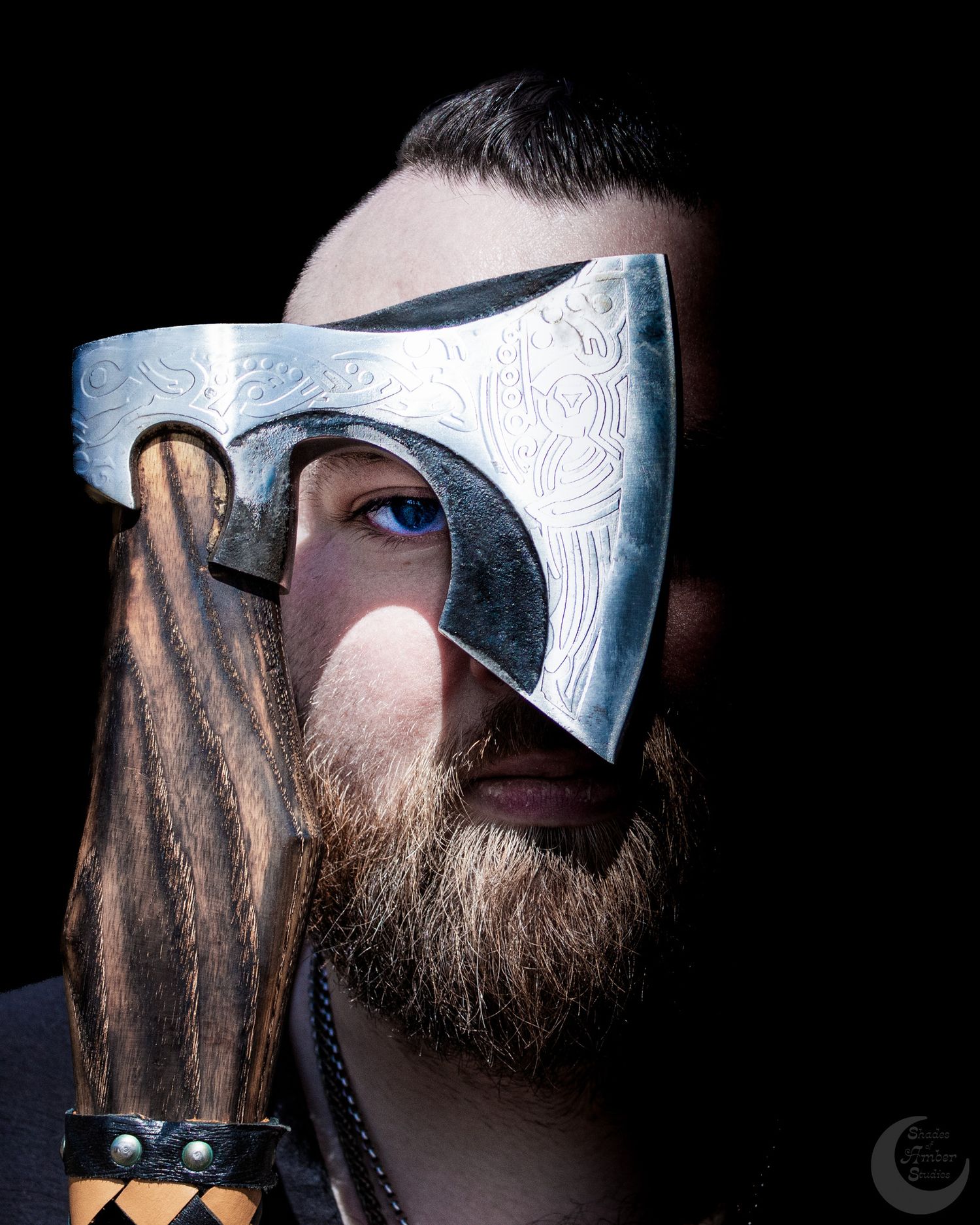 Bearded man holding an engraved Viking axe up to his face against a dark background.