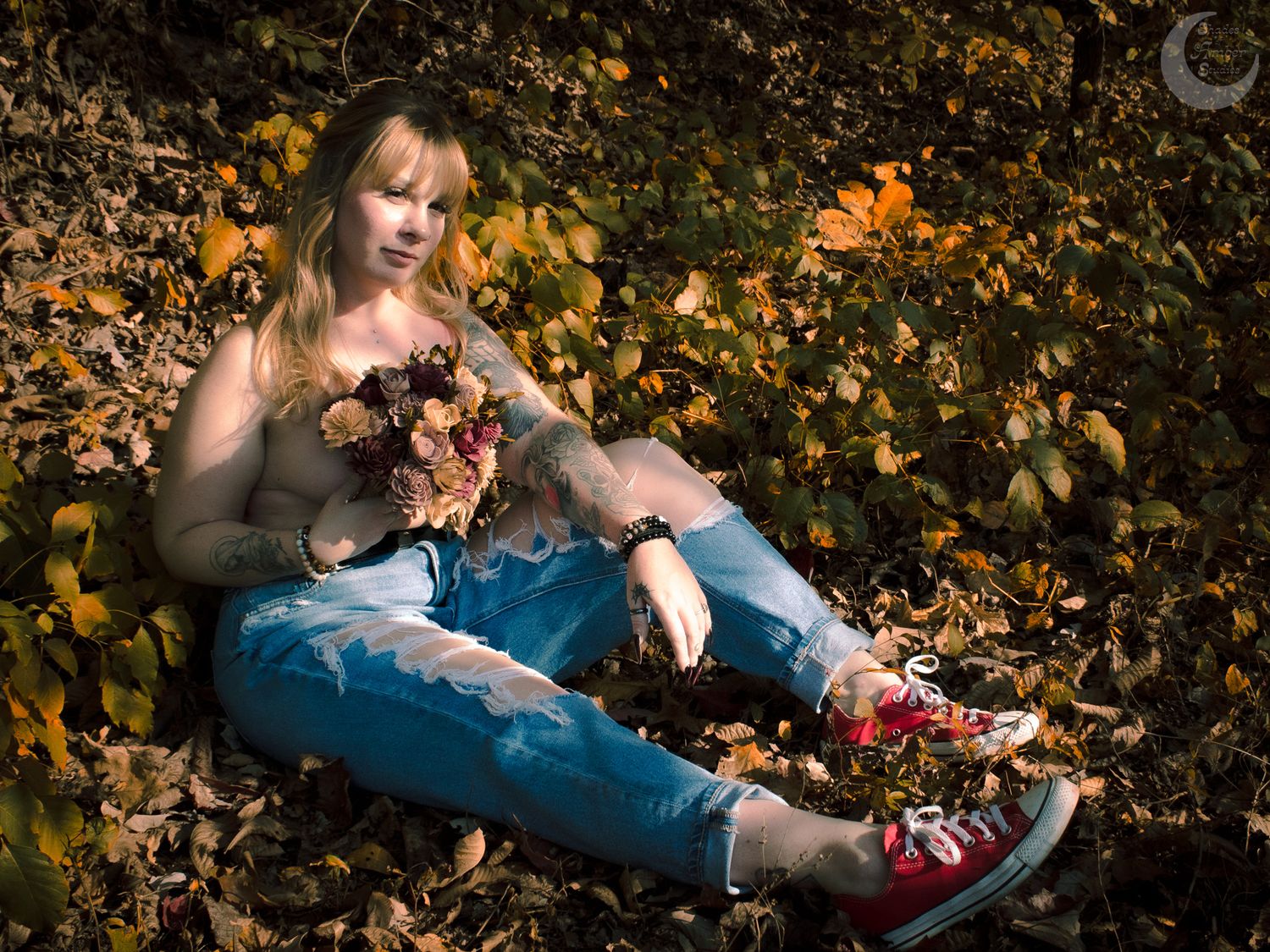 Person sitting among autumn leaves wearing jeans and holding autumn foliage in moody outdoor portrait.
