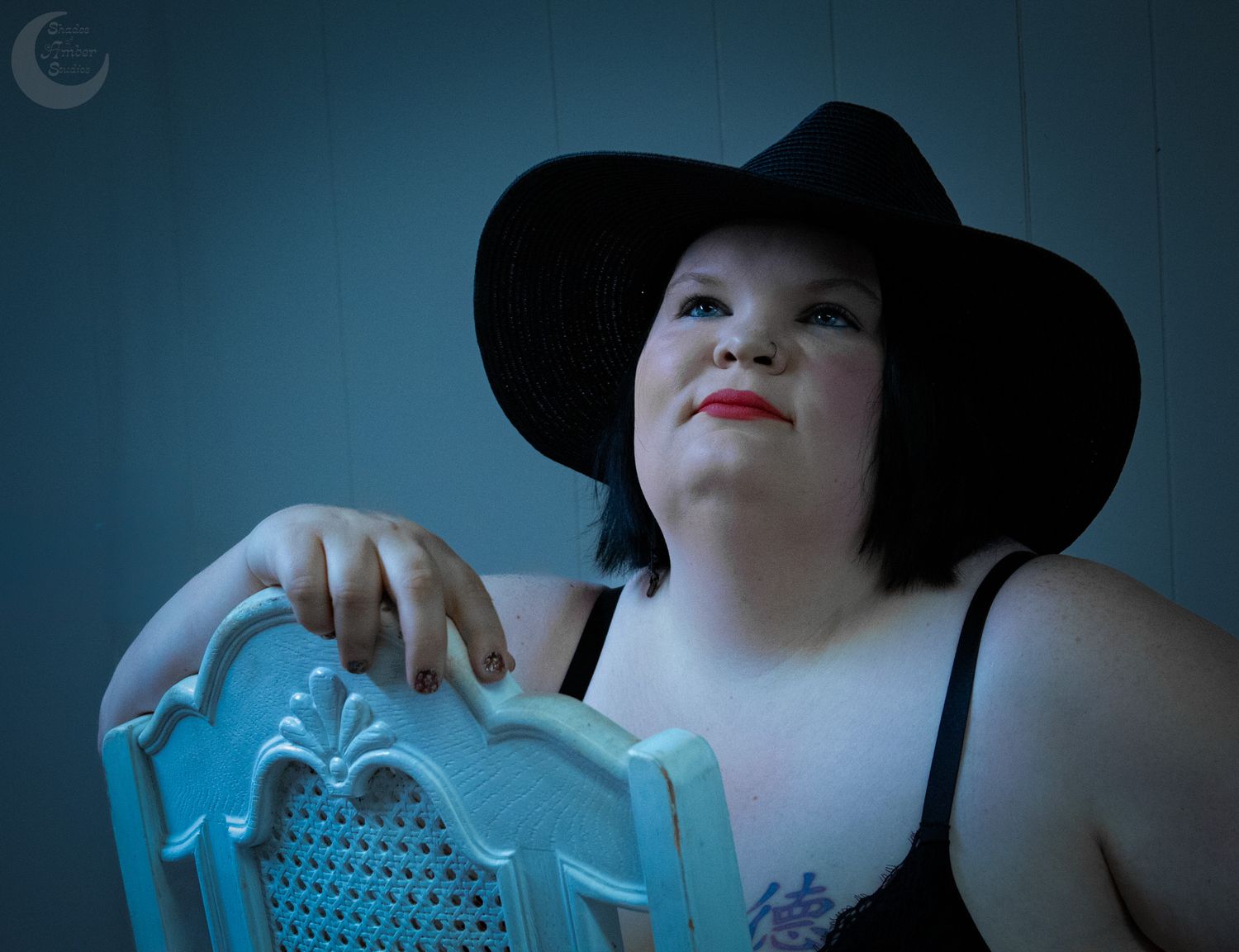 Someone in a black hat poses with a turquoise vintage chair against a dark background in dramatic lighting.