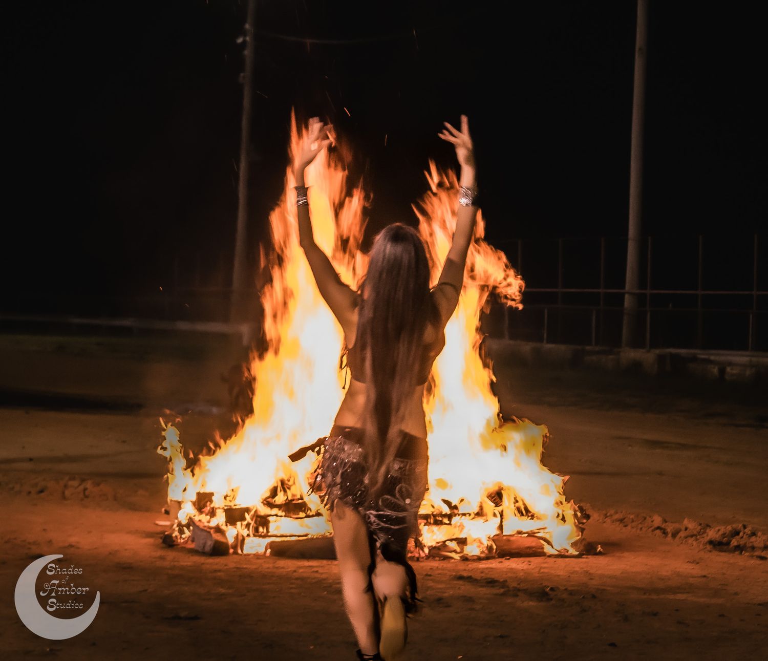 A person stands in front of a large bonfire at night with arms raised in a dramatic pose.