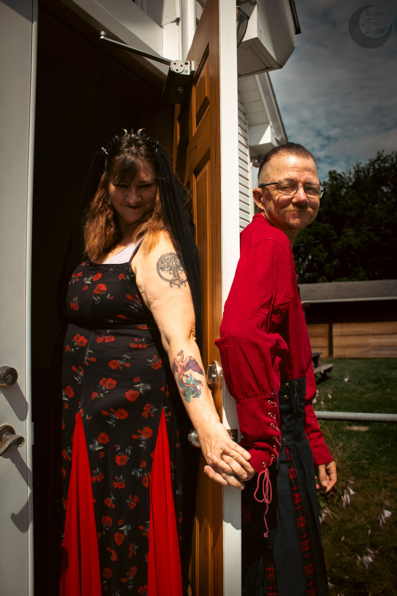 A couple in coordinated red and black gothic-inspired outfits stand in a doorway holding hands.