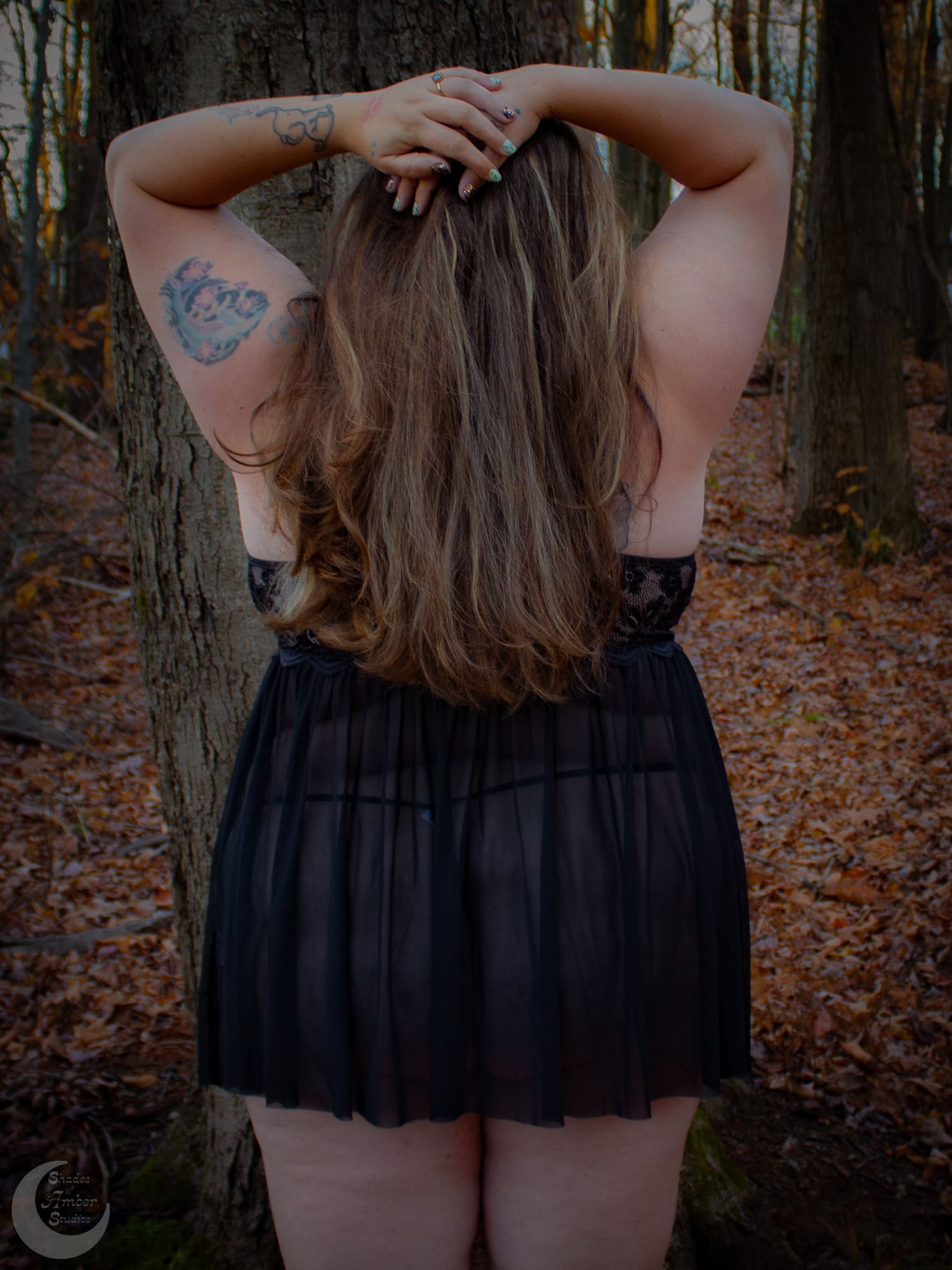 A back view portrait showing long hair and tattoos in a sheer black dress among fall foliage.