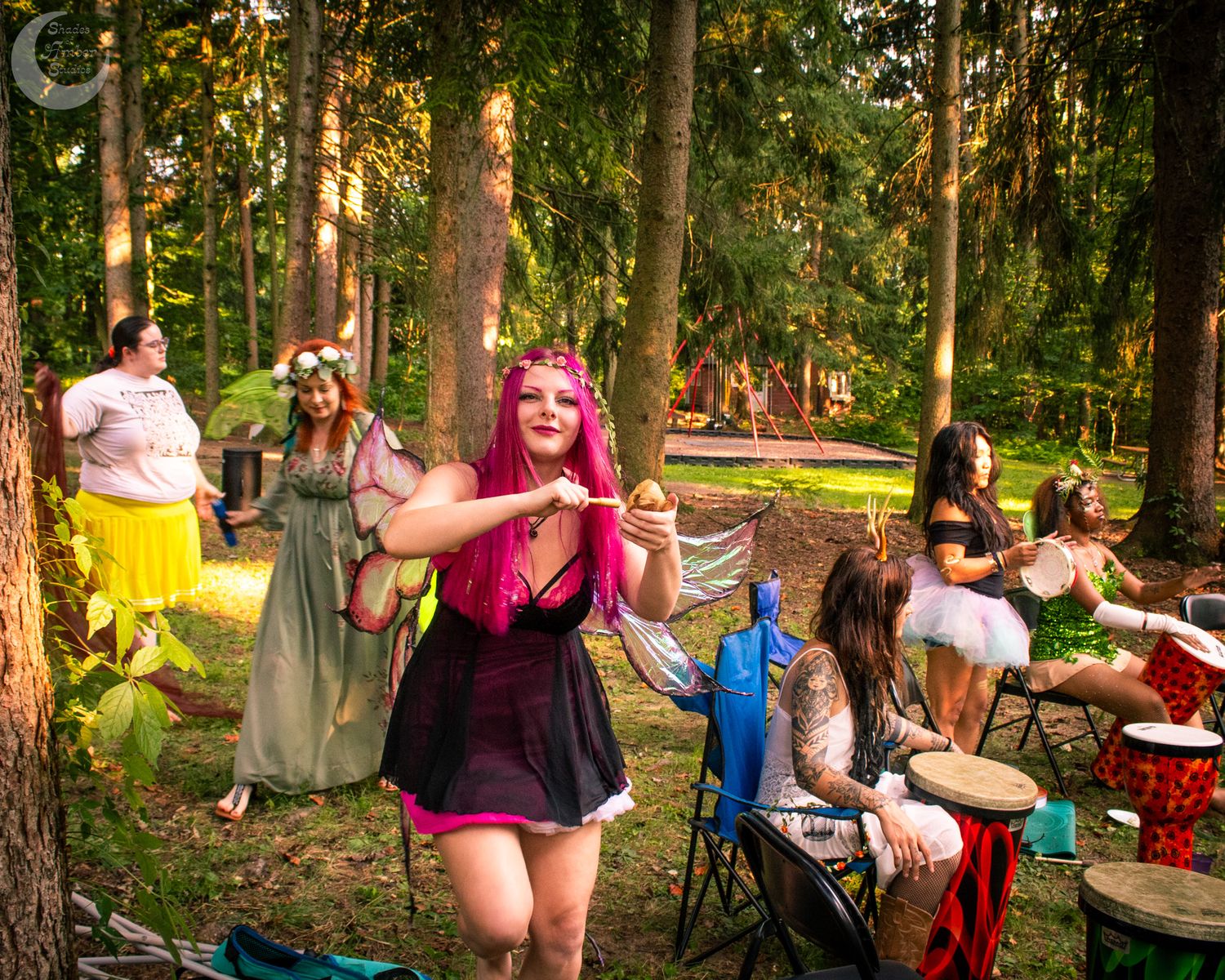 Festival attendees gather for an outdoor summer party in a wooded setting with fairy lights and colorful decorations.