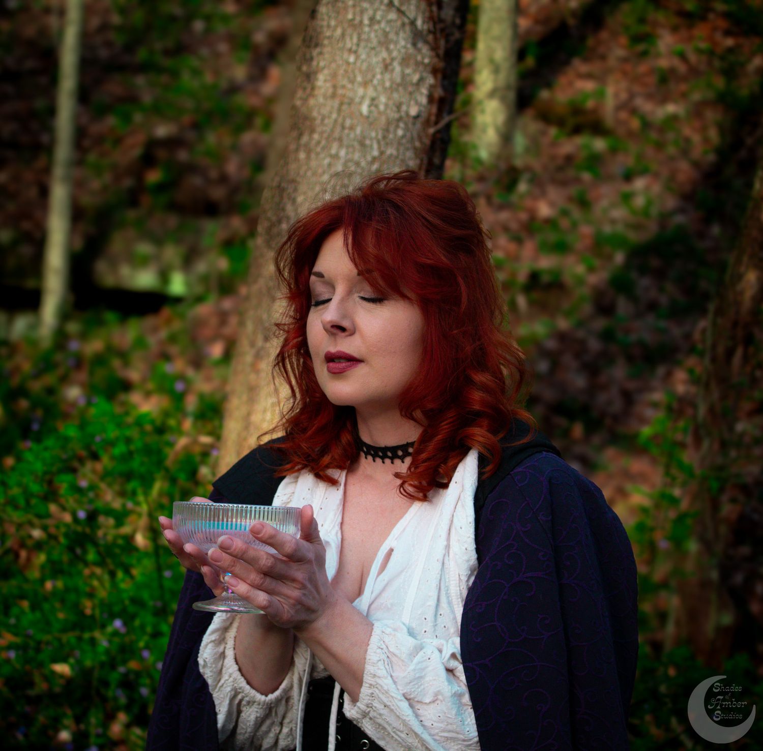 A person with red hair writes in a notebook while sitting in a wooded area wearing a white blouse and navy cardigan.
