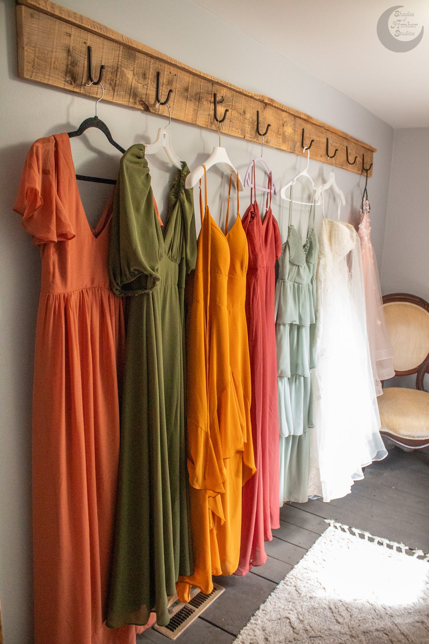 Colorful bridesmaid dresses in rust, olive green, gold and mint hang on rustic wooden hooks mounted on a light wall.