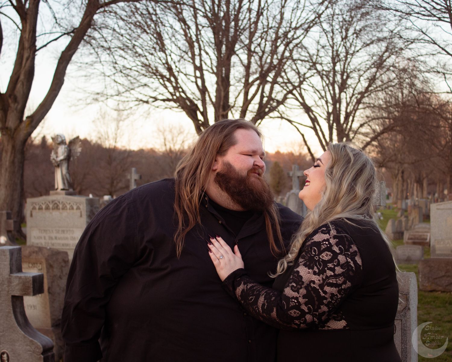 Couple in black attire sharing a romantic moment in a cemetery with bare trees at sunset.