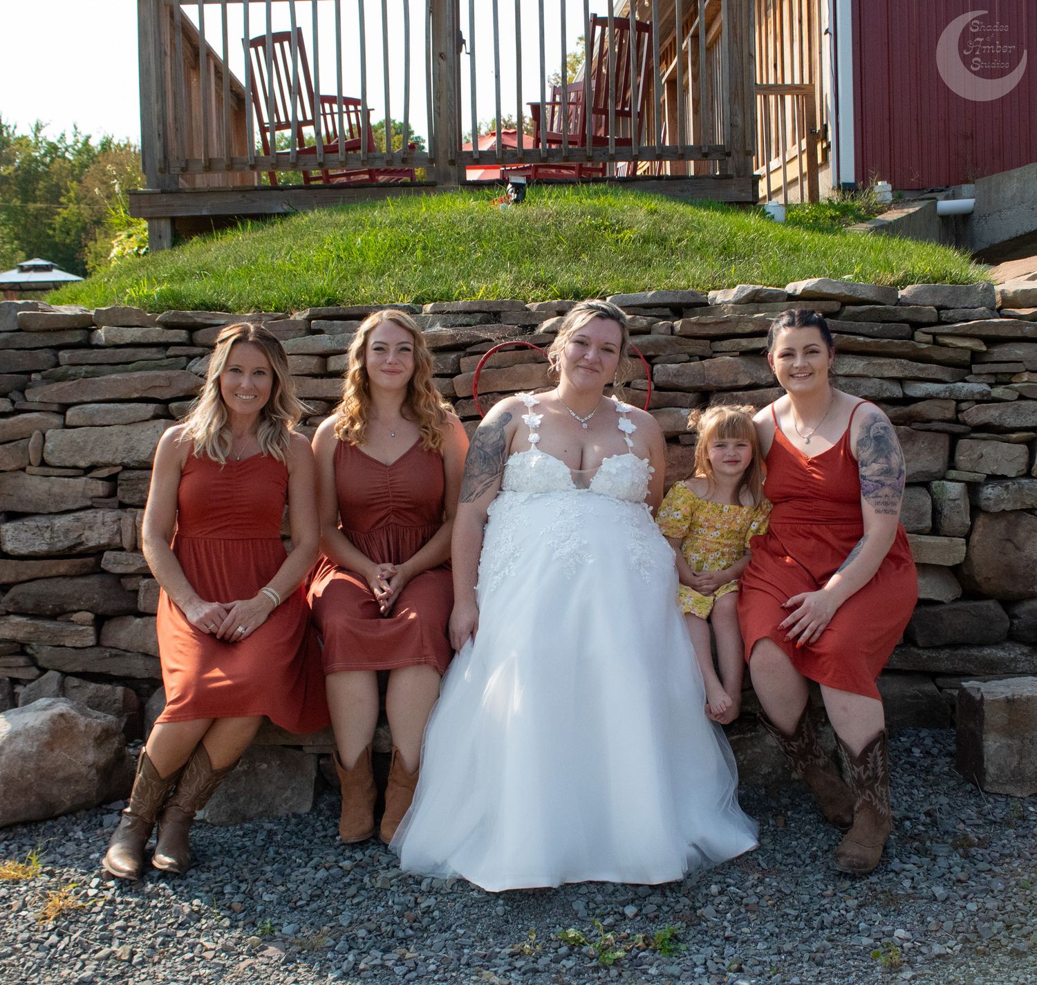 Bridal party in rustic orange dresses sitting on stone wall outside barn.