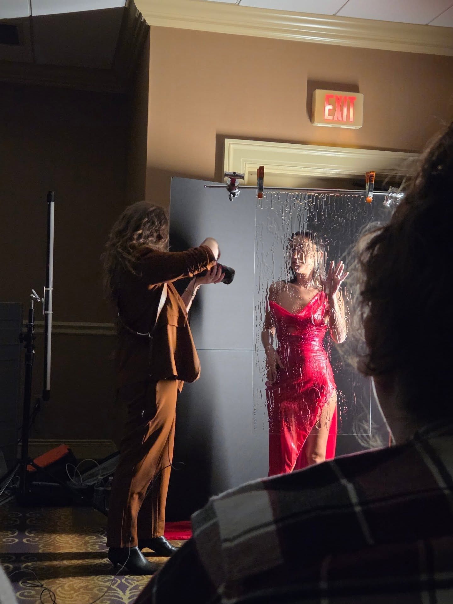 A photographer captures a model in a red dress during a professional photoshoot in a hallway setting.