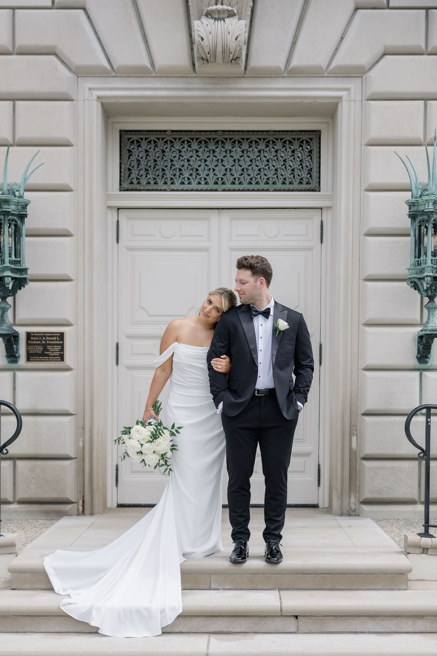 Bride in off-shoulder gown and groom in tuxedo pose on steps before elegant white building doors.