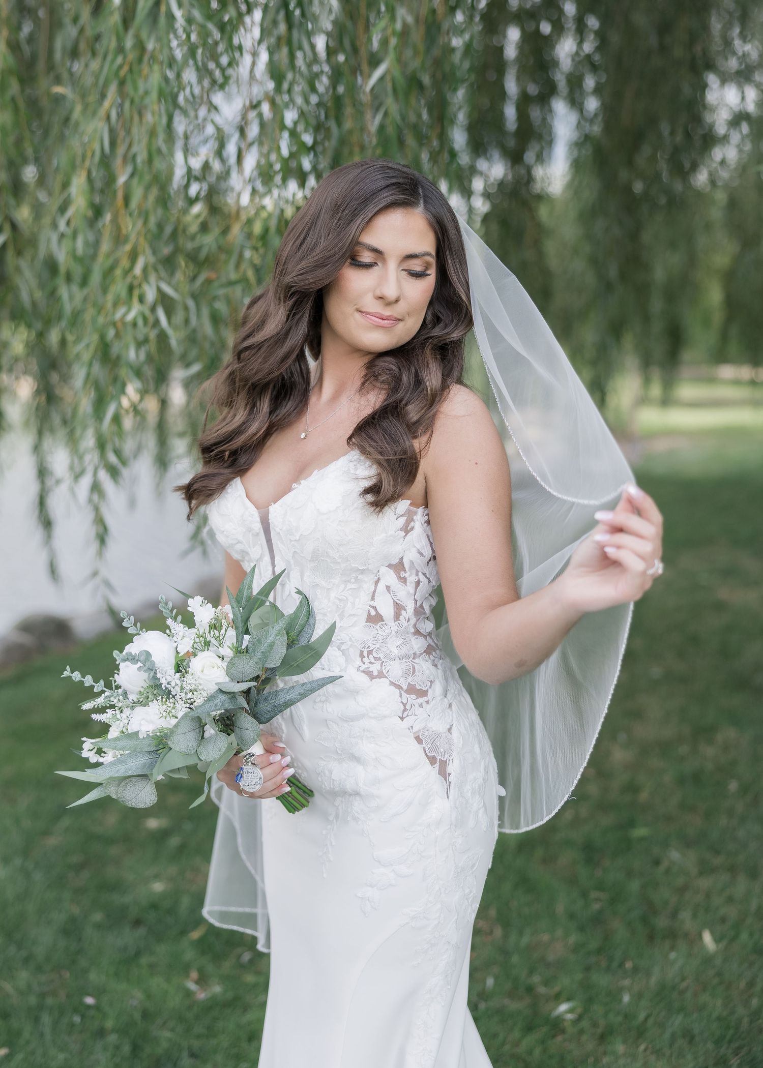 A bride in a fitted lace wedding gown holds a eucalyptus bouquet while standing beneath a willow tree.