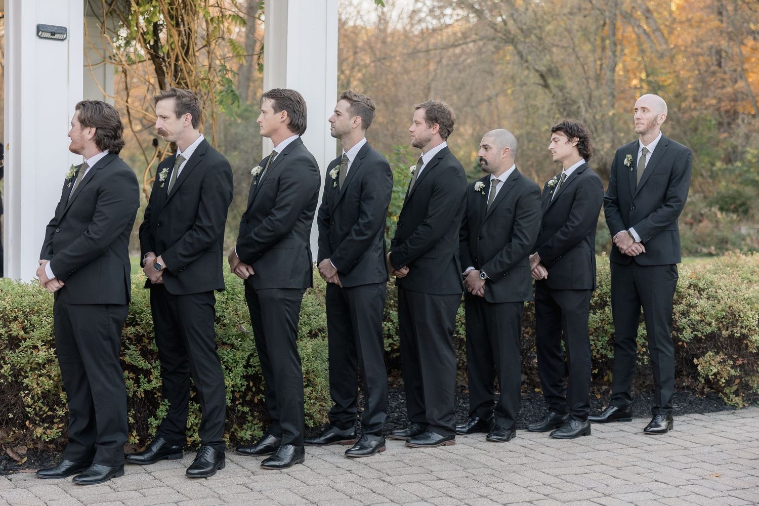 Groomsmen in black suits lined up at an outdoor wedding ceremony.