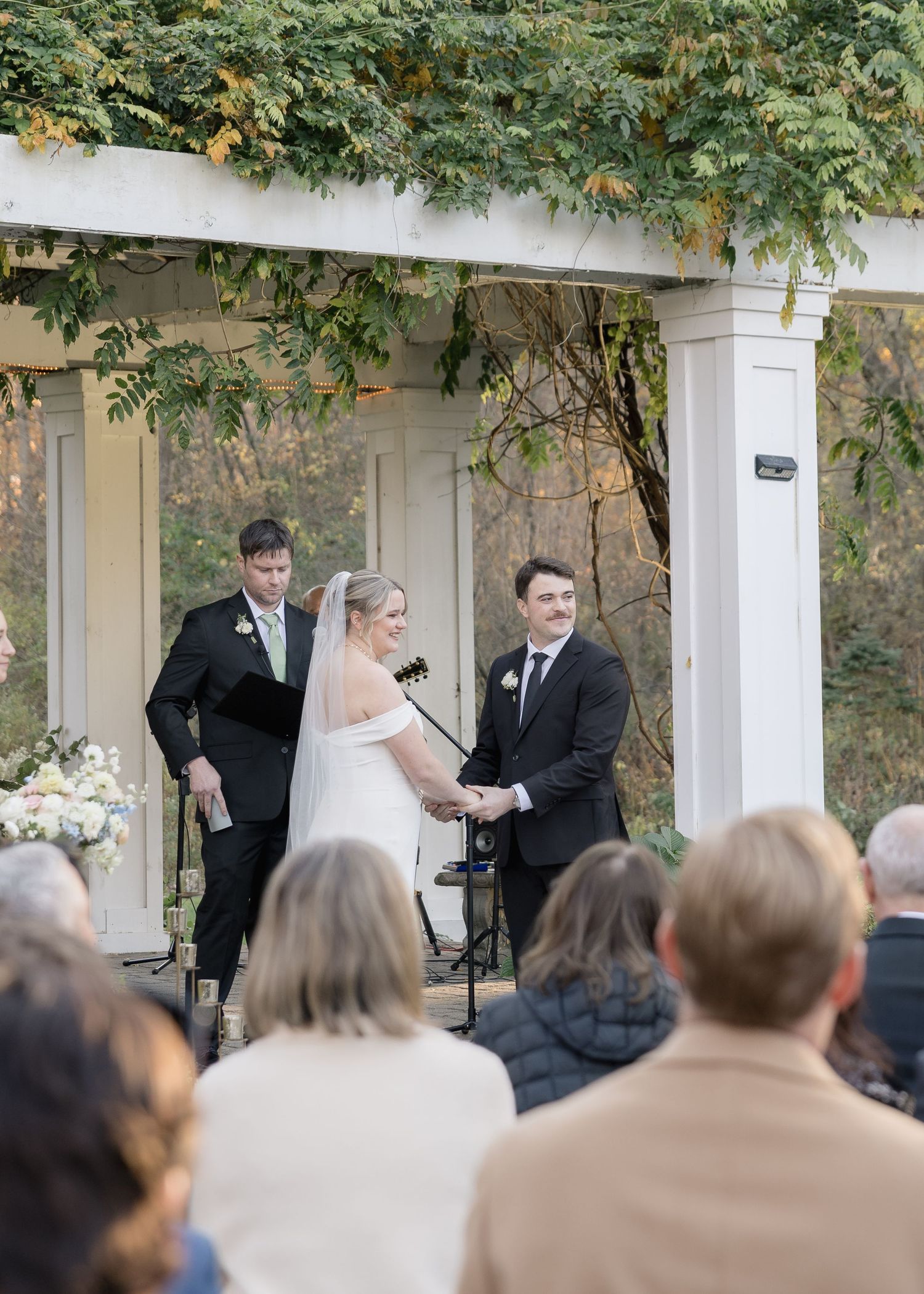 Garden wedding ceremony with guests seated beneath pergola decorated with climbing vines.
