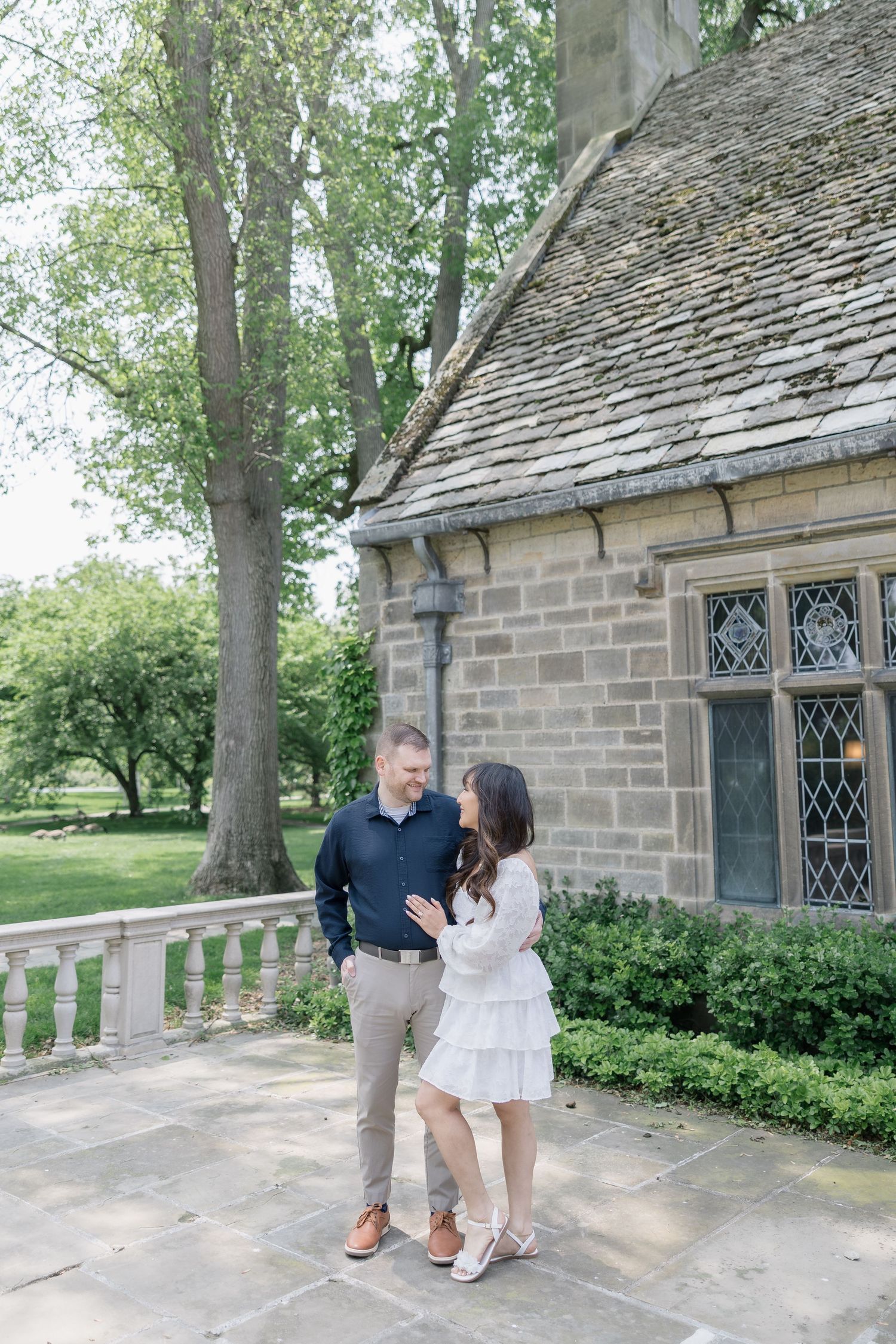 A couple poses in front of a historic stone chapel building in a park-like setting.