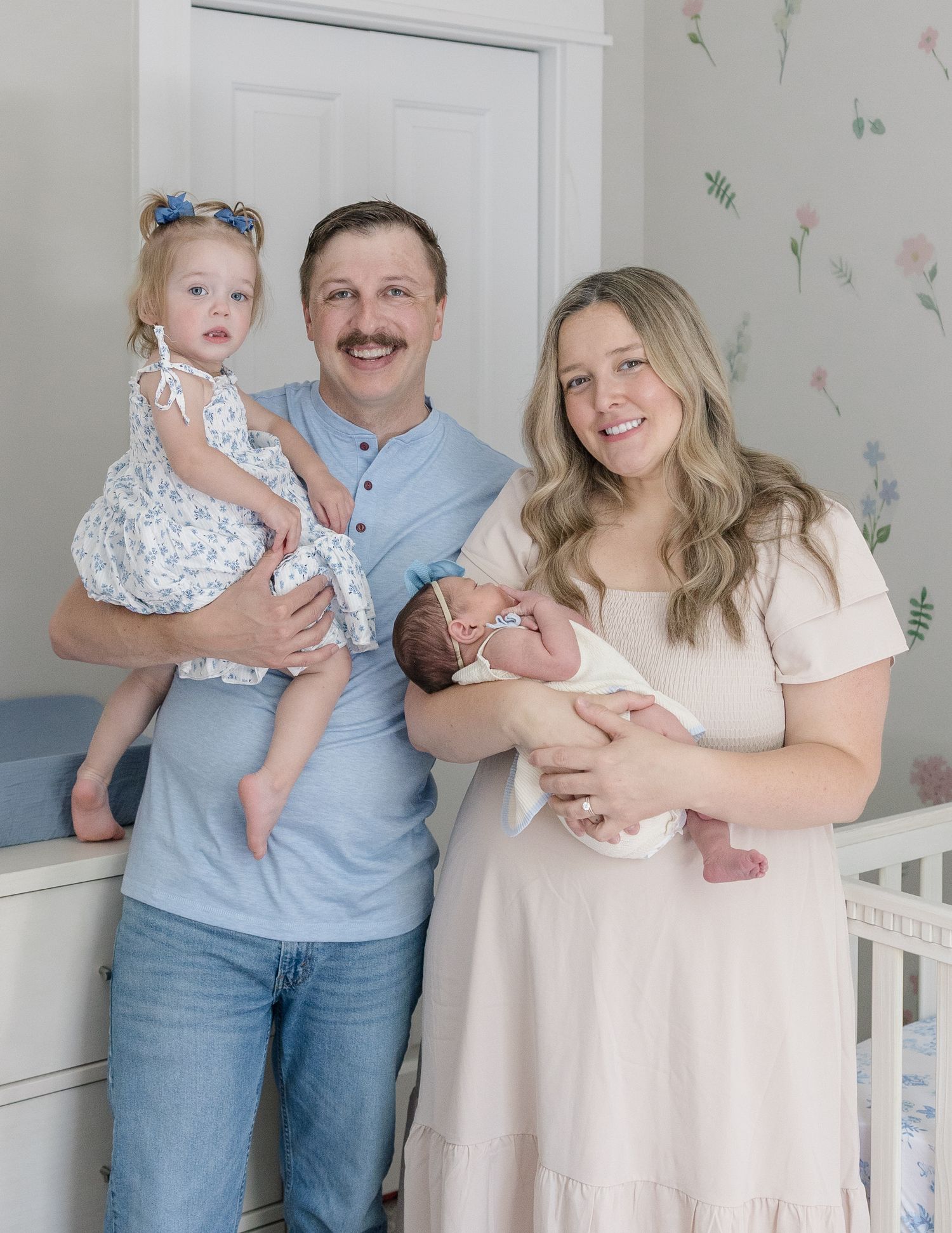 A happy family of four poses in a nursery, with dad holding a toddler girl and mom cradling a newborn baby.