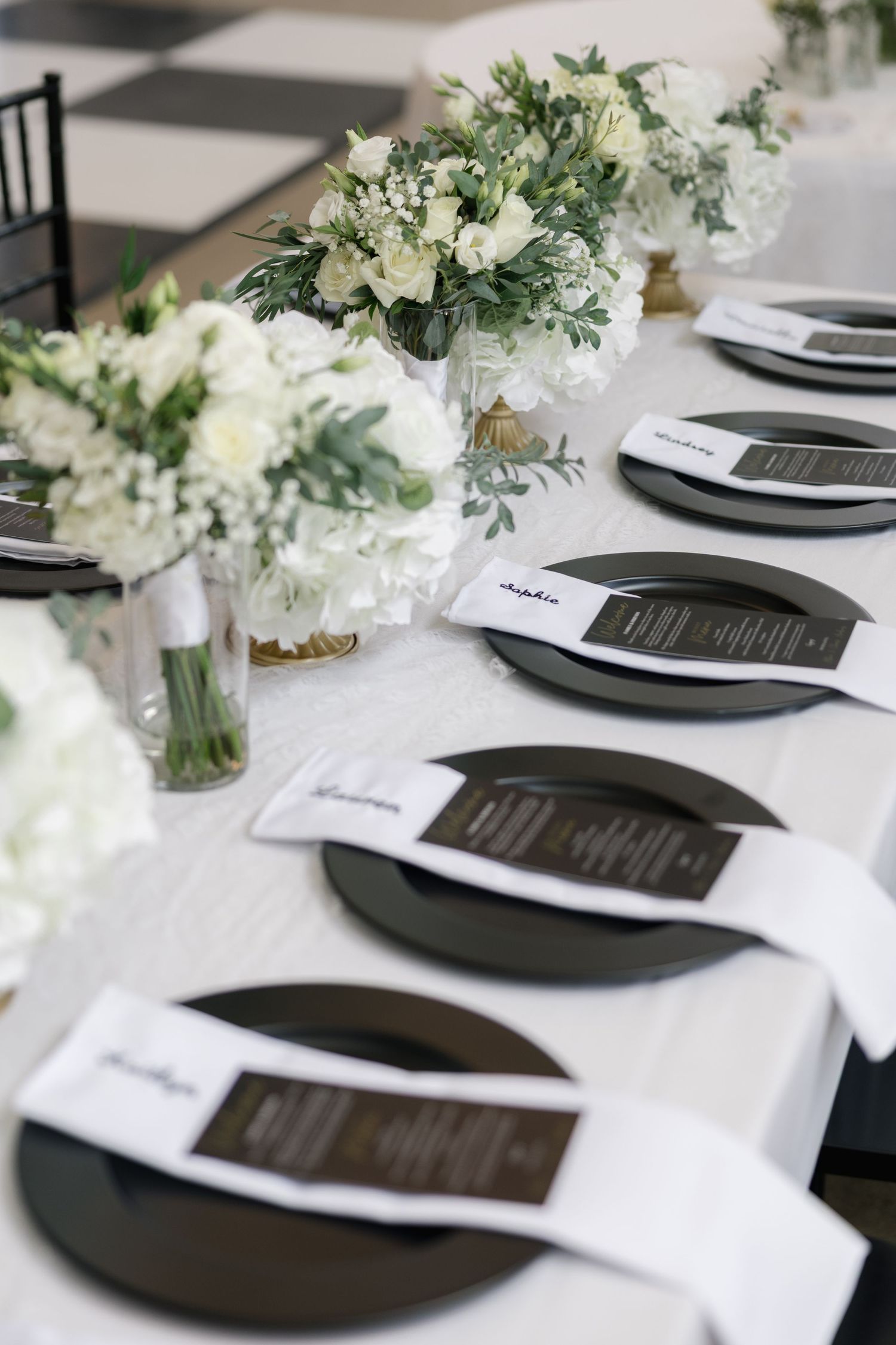 Elegant wedding table setting with black plates, white menus, and white floral centerpieces on a white linen tablecloth.