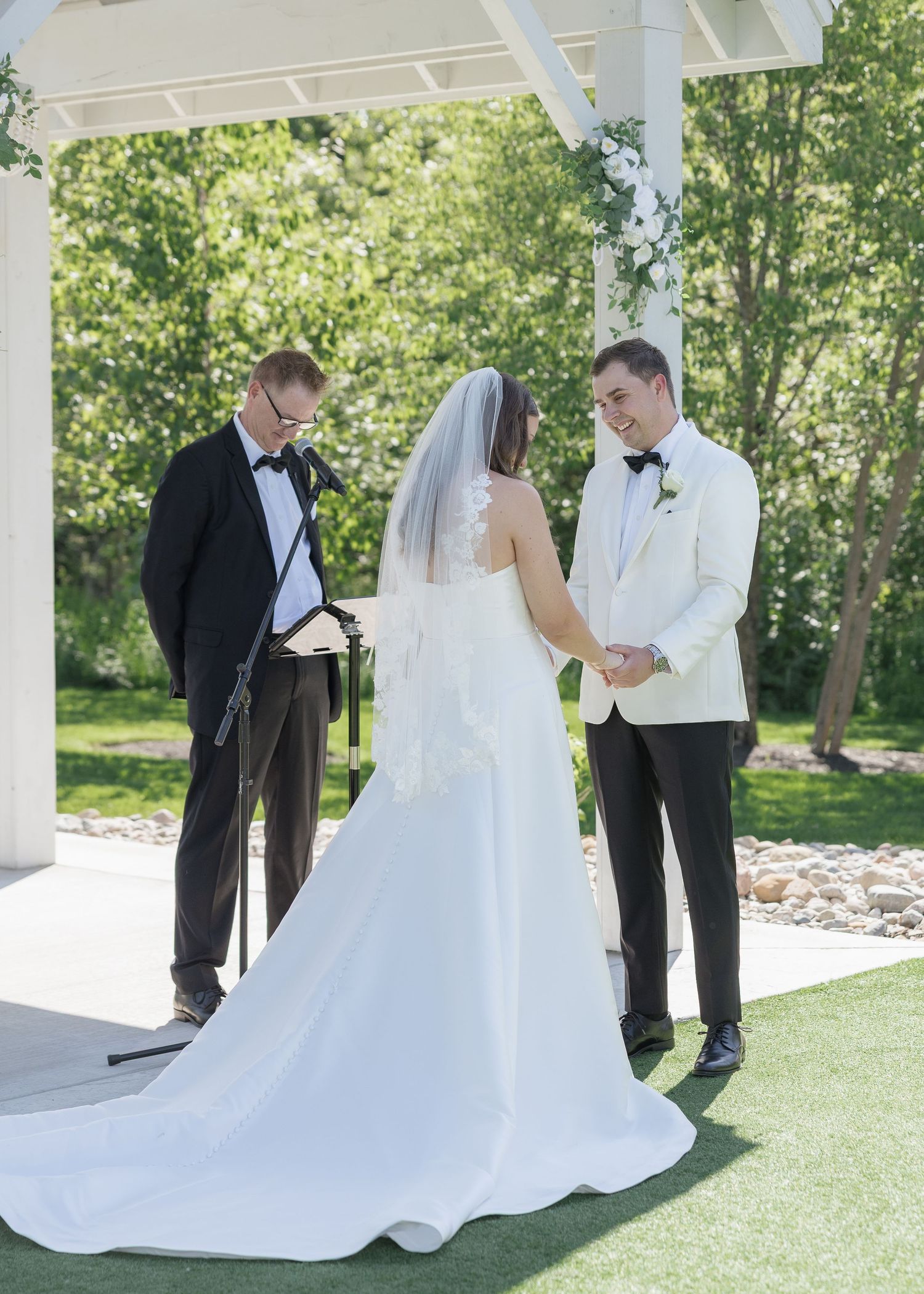 Bride and groom exchange vows outdoors under a white pergola, officiant reading from a book, lush greenery background.
