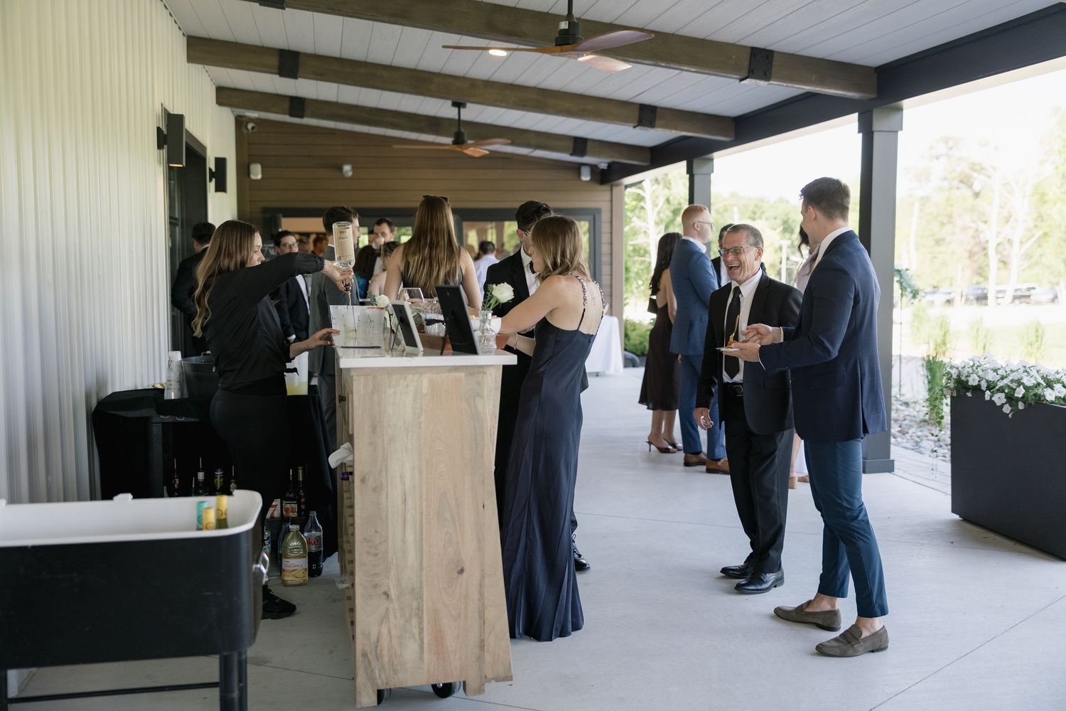 Guests mingle at an elegant outdoor cocktail hour under a covered patio with a rustic wooden bar.