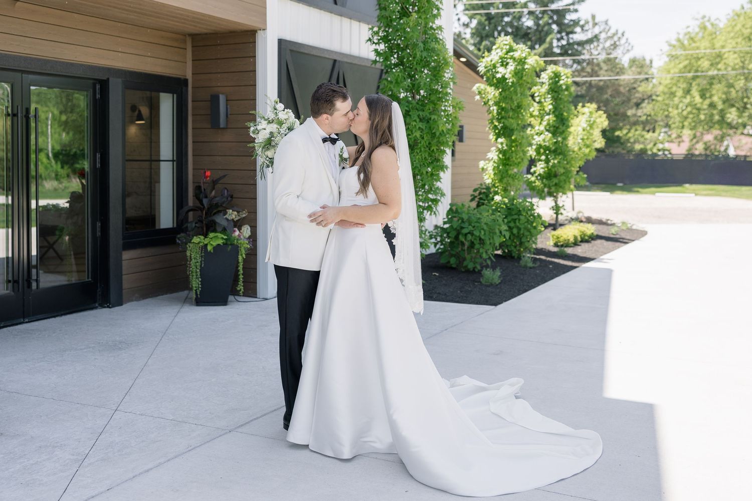A bride and groom share a romantic kiss outside a modern venue, surrounded by lush greenery on their wedding day.