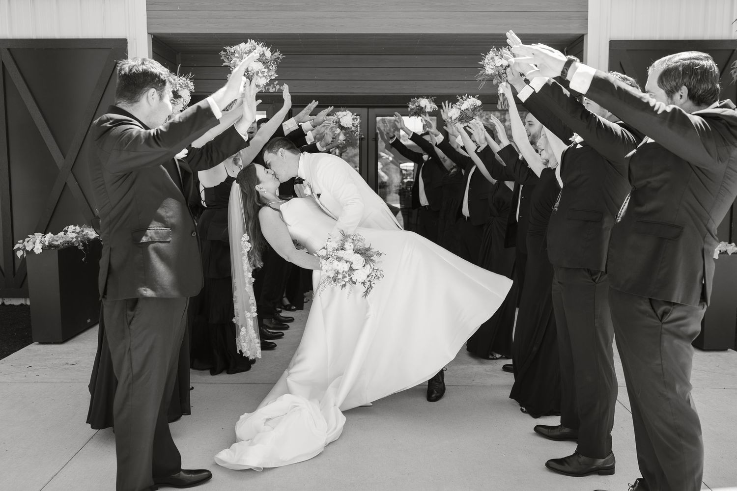 Black and white photo of bride and groom sharing romantic dip kiss under archway.