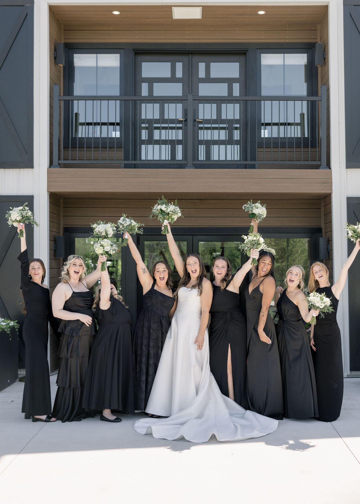 Bride in white gown celebrates with bridesmaids in black dresses raising bouquets outside modern venue.