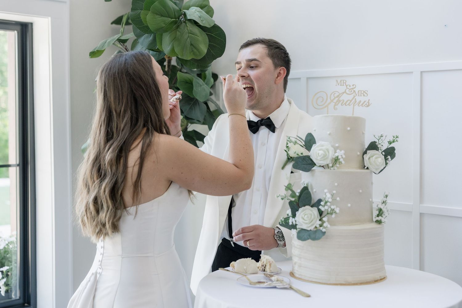 Bride playfully feeds groom a piece of wedding cake during their reception near a white floral tiered cake.