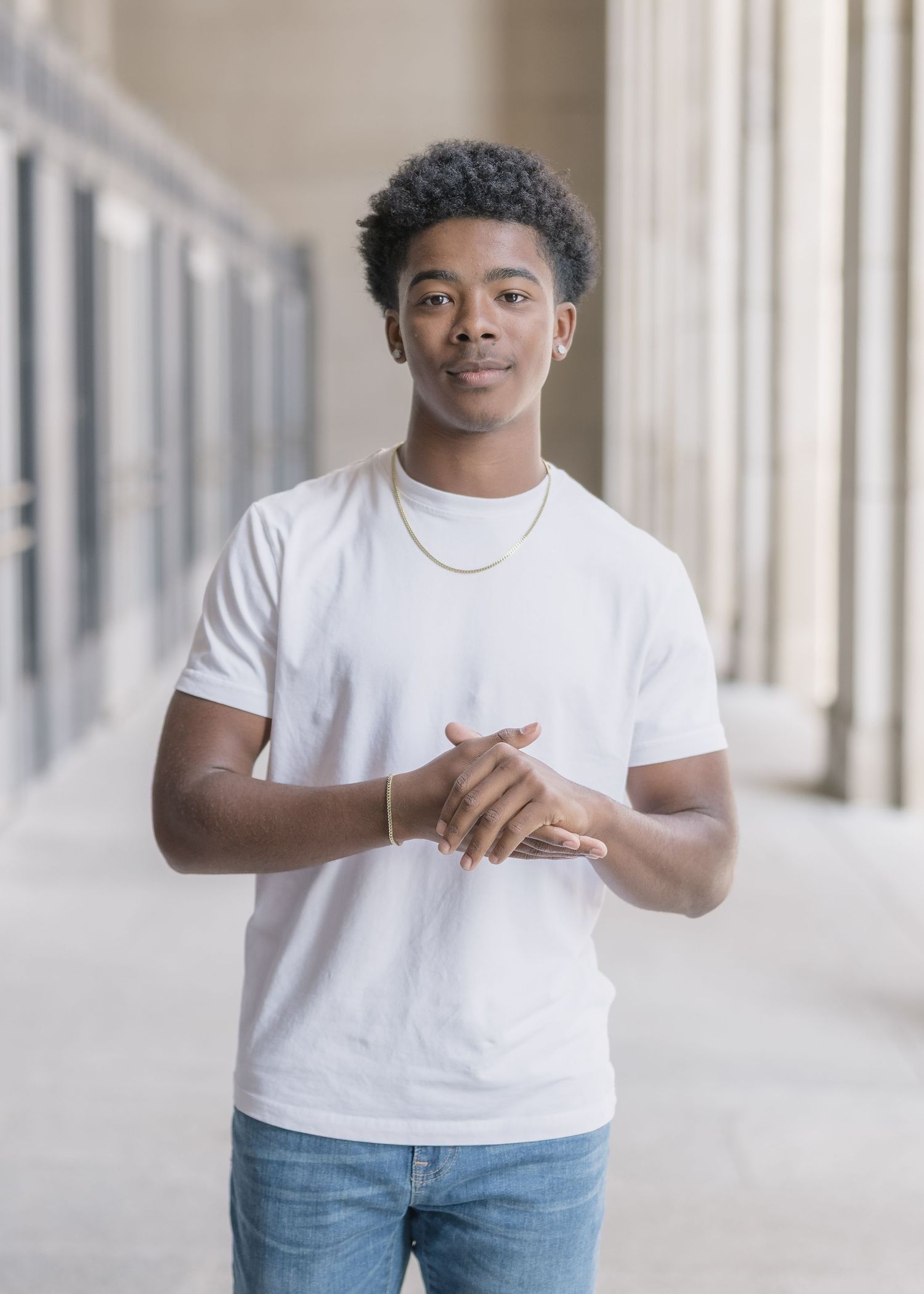 Young person in a white t-shirt and jeans stands confidently in a hallway with columns while wearing a gold necklace.
