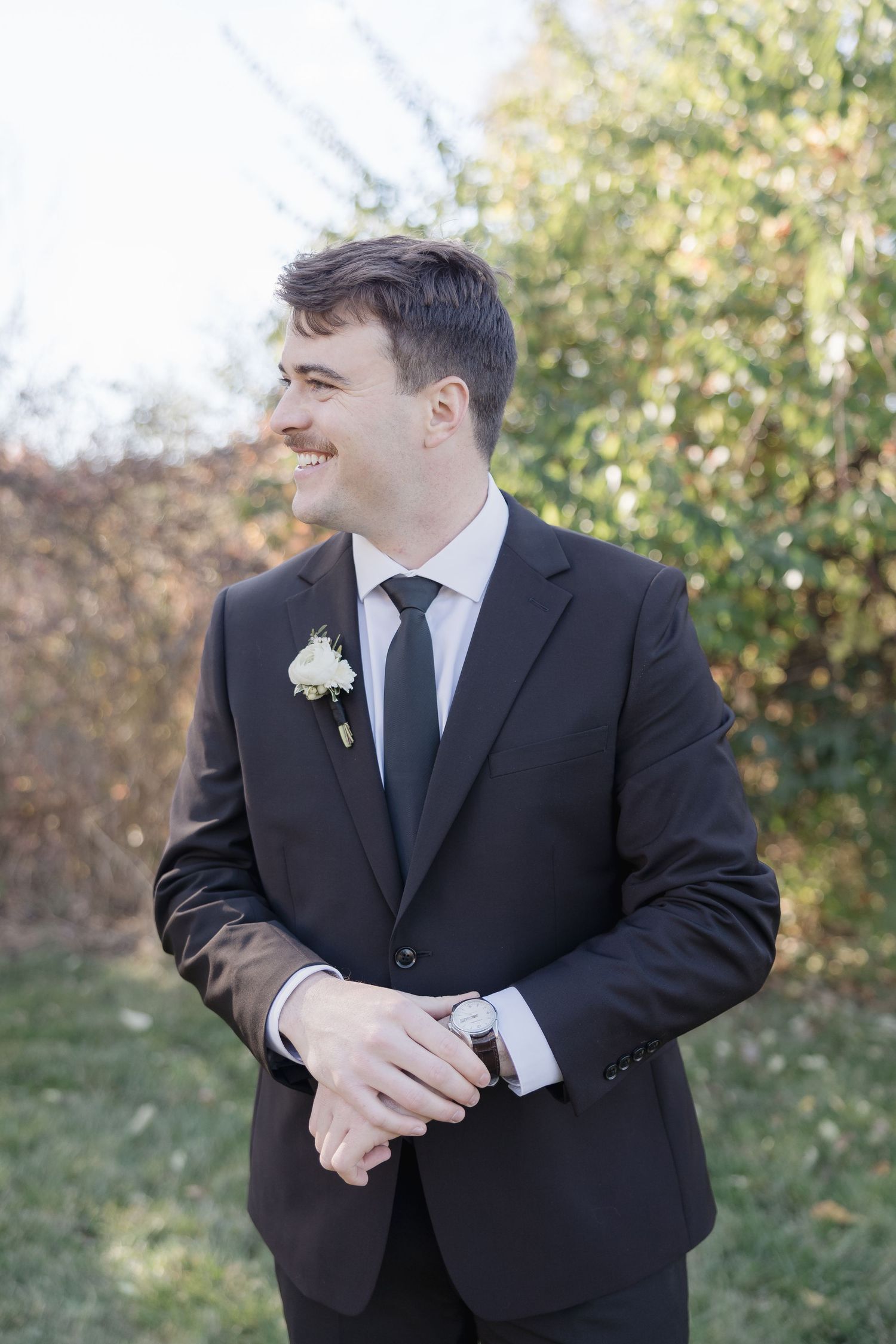 Groom in classic black suit and tie smiles while adjusting his watch against autumn foliage background.