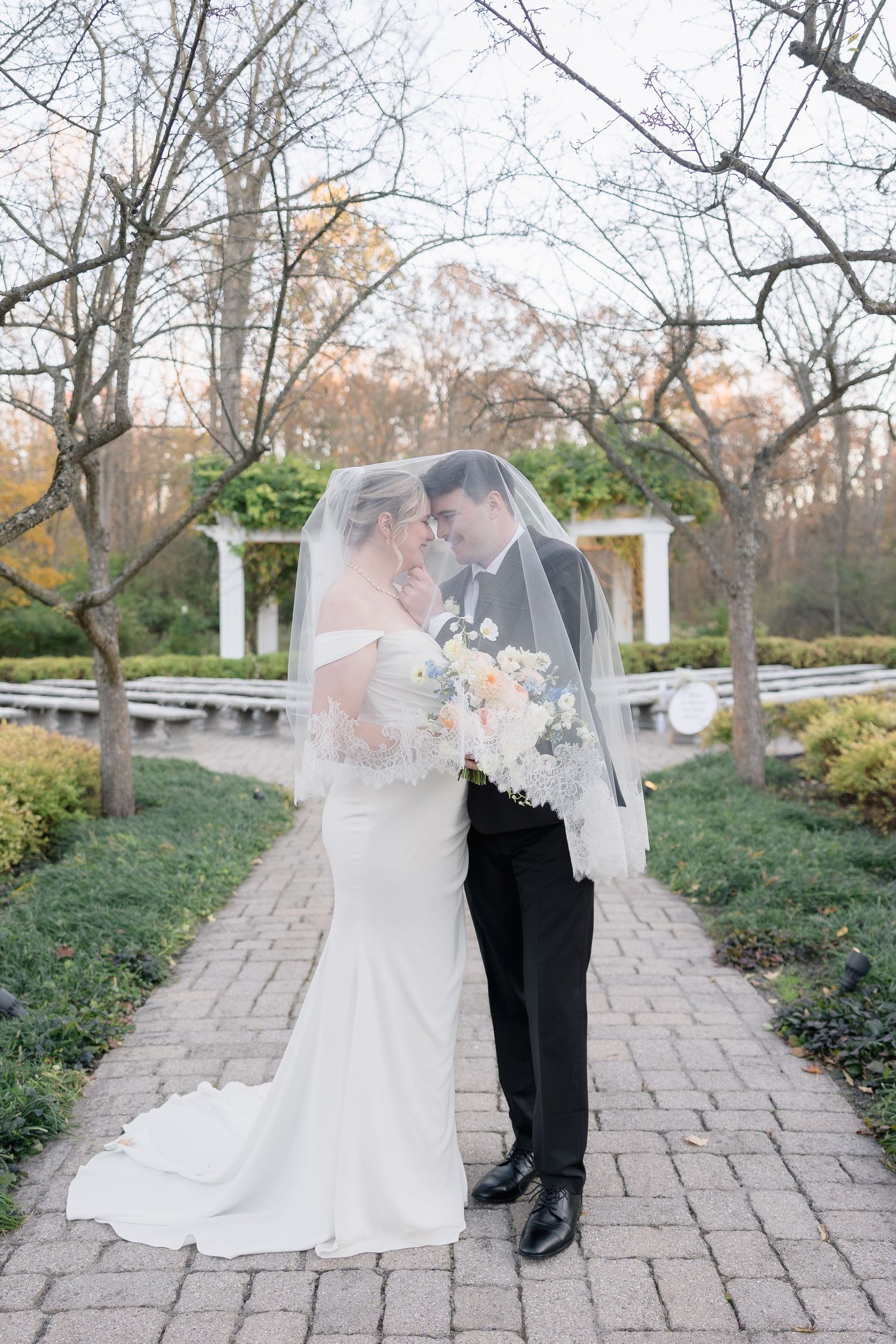 Ethereal wedding portrait with couples embracing under flowing veil on garden path.
