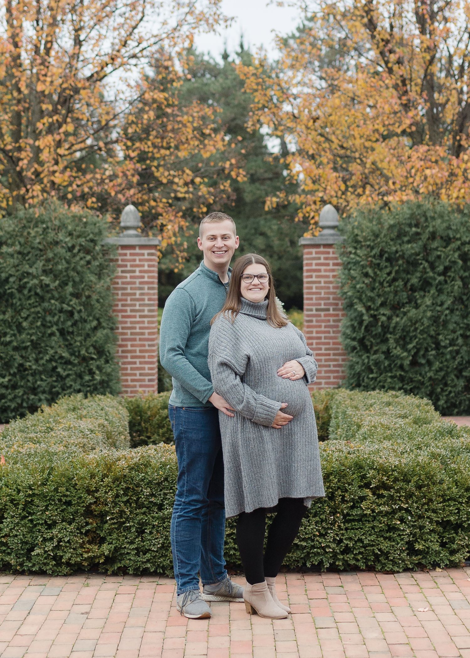 Maternity portrait of couple standing on brick pathway with autumn trees and brick pillars.