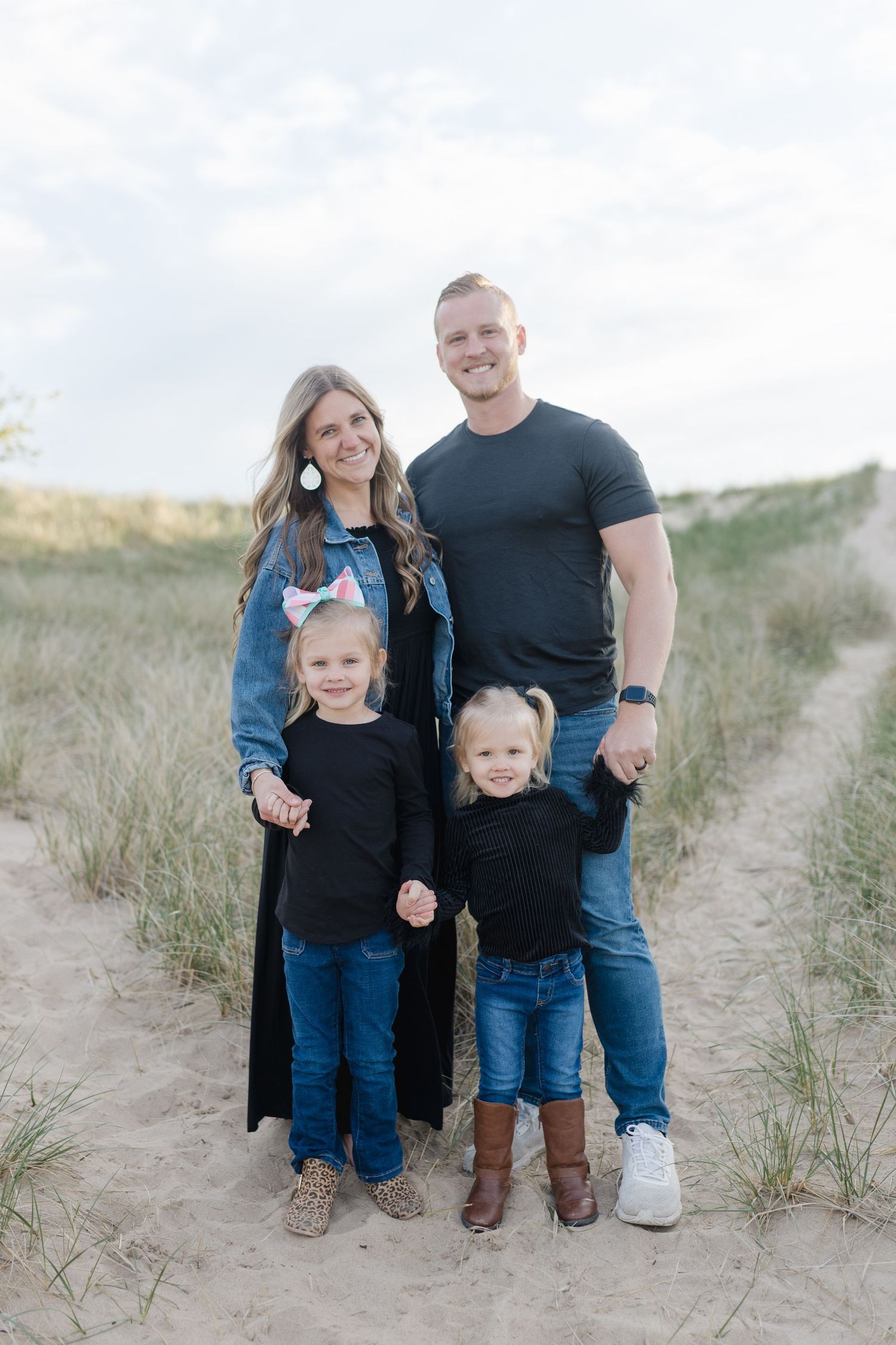 A family wearing coordinating blue and black outfits poses together on sandy dunes at the beach.