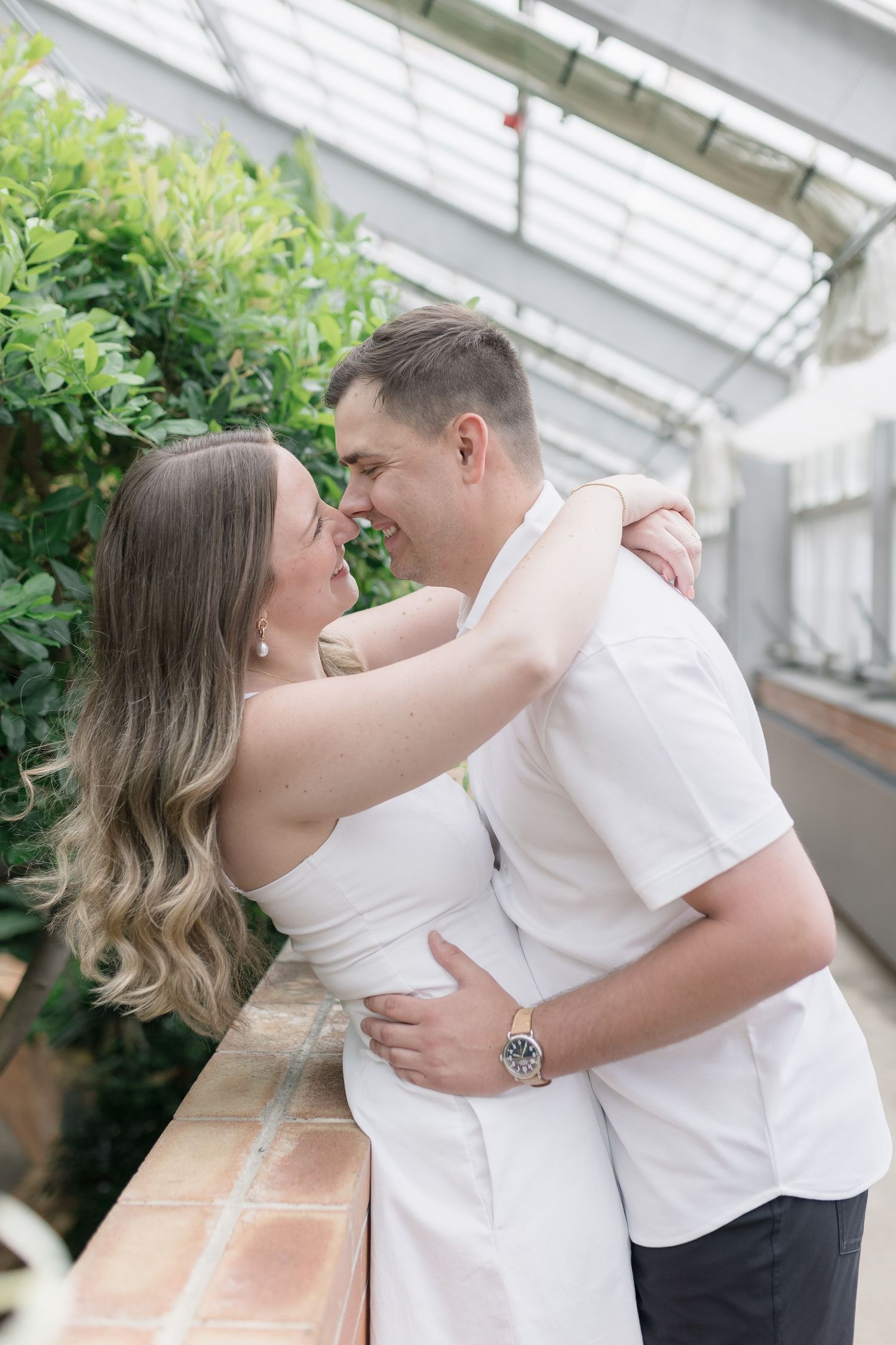 A romantic couple embraces in a greenhouse setting with lush greenery and white walls in the background.
