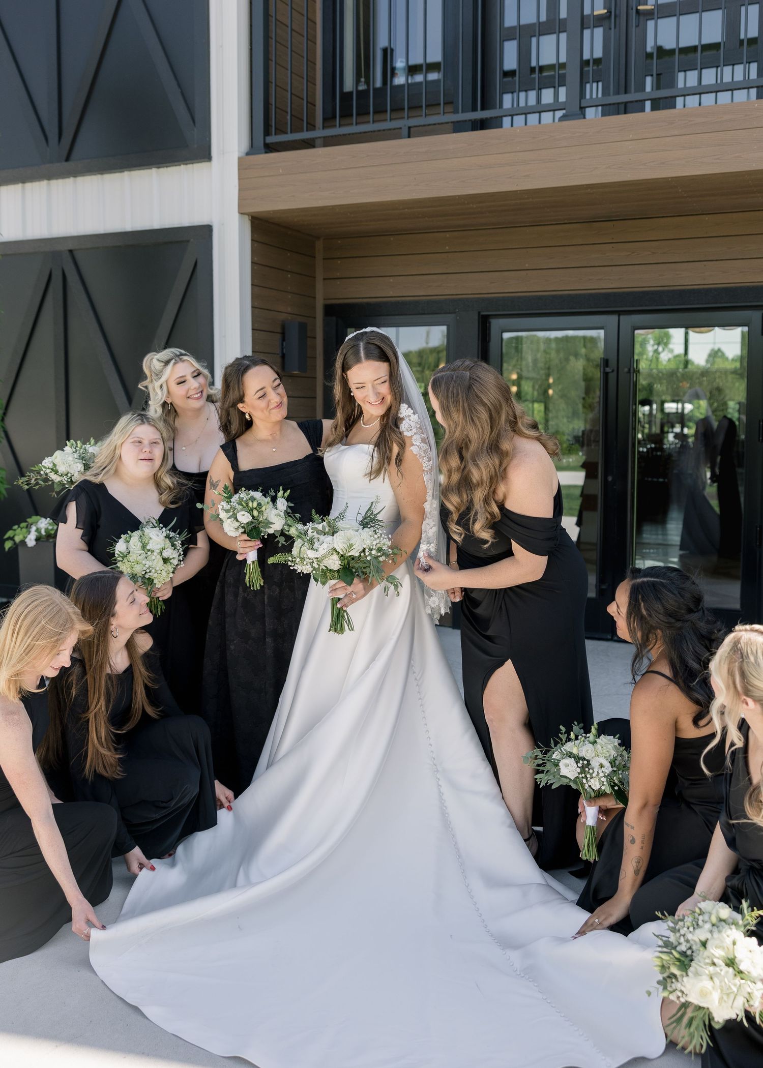 Bride in white gown surrounded by bridesmaids in black dresses holding white floral bouquets outdoors.