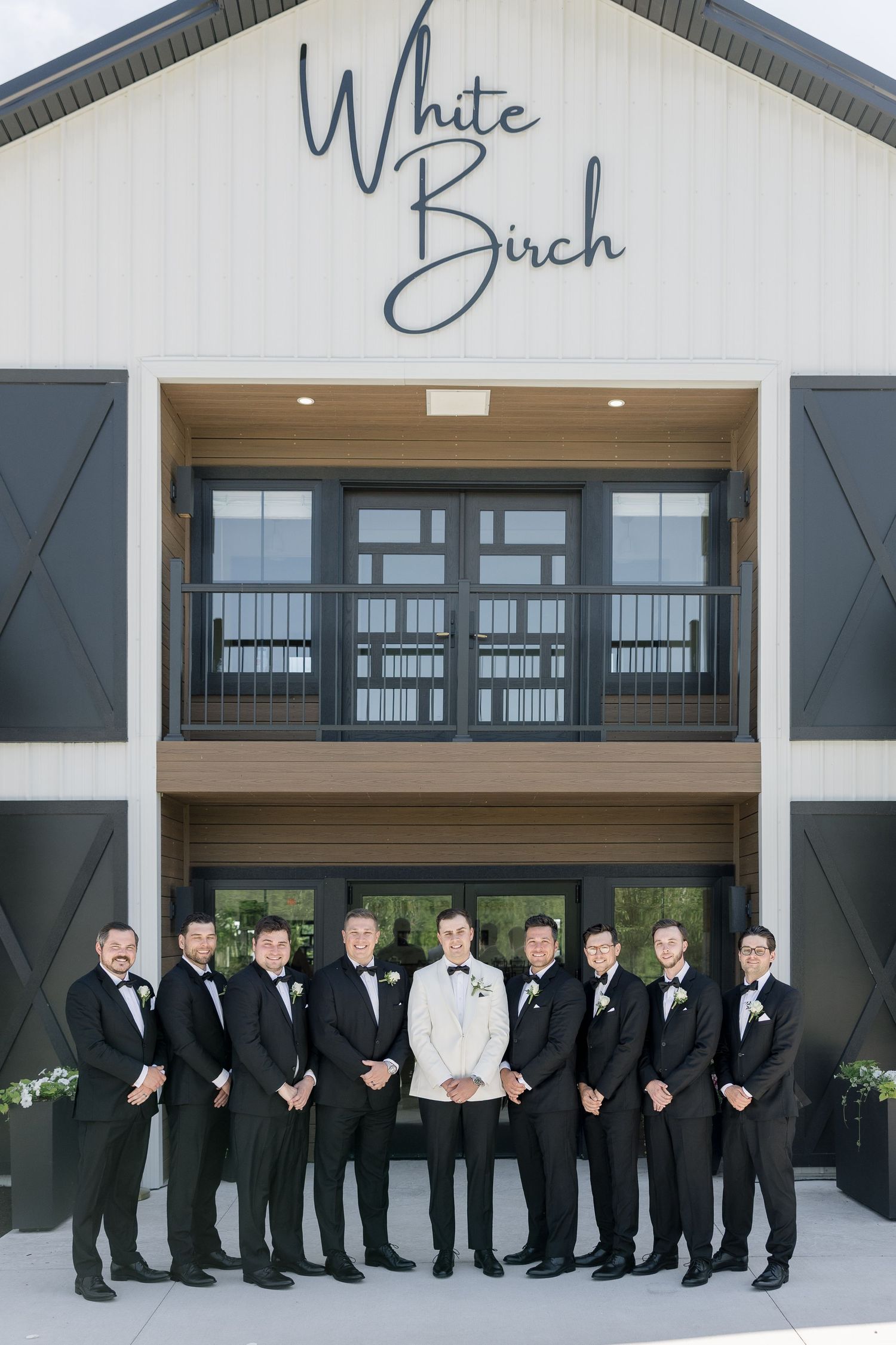 Groom and groomsmen in black tuxedos pose outside White Birch wedding venue with modern barn architecture.