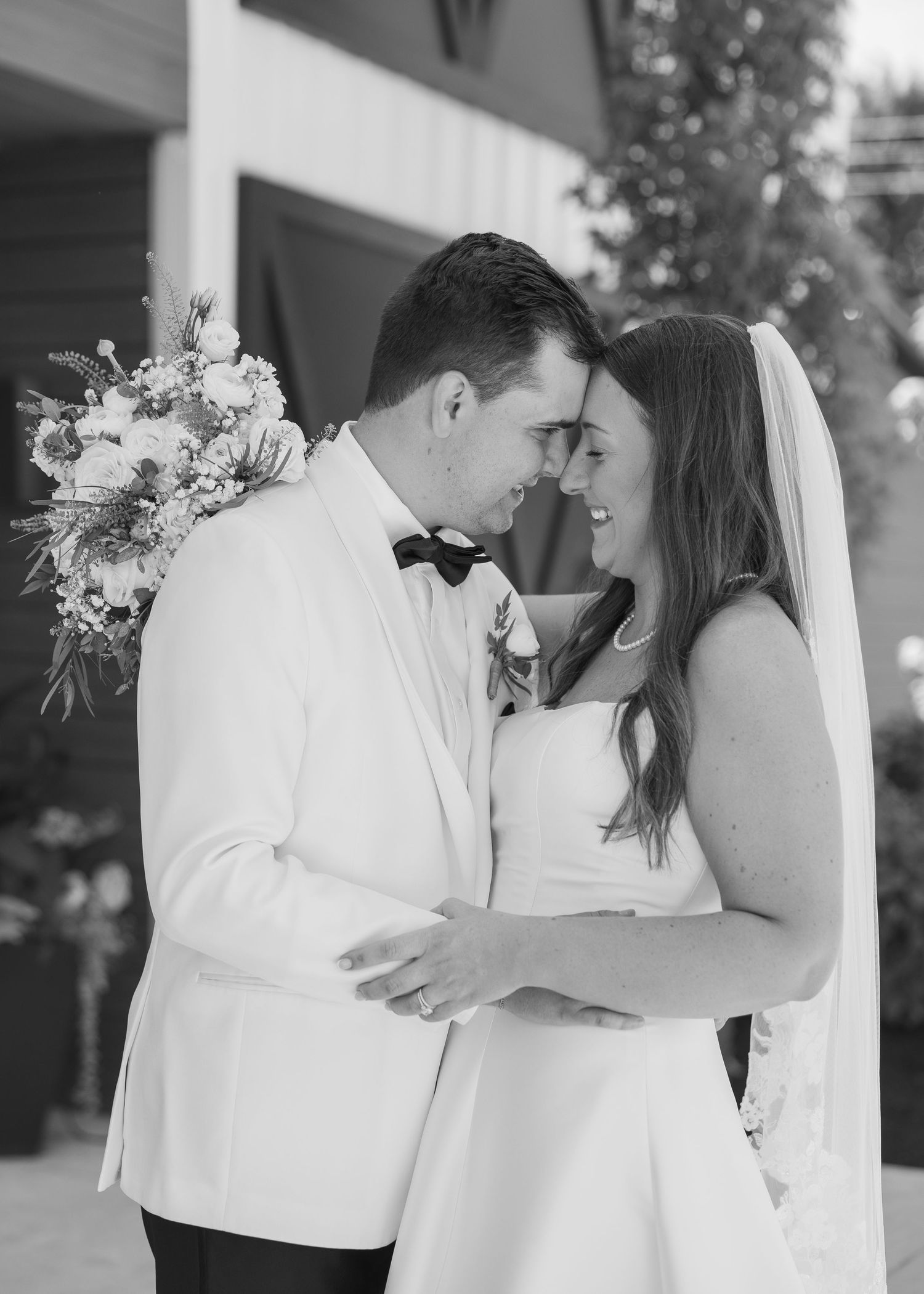 Black and white photo of bride and groom touching foreheads, smiling intimately, bride holding floral bouquet.