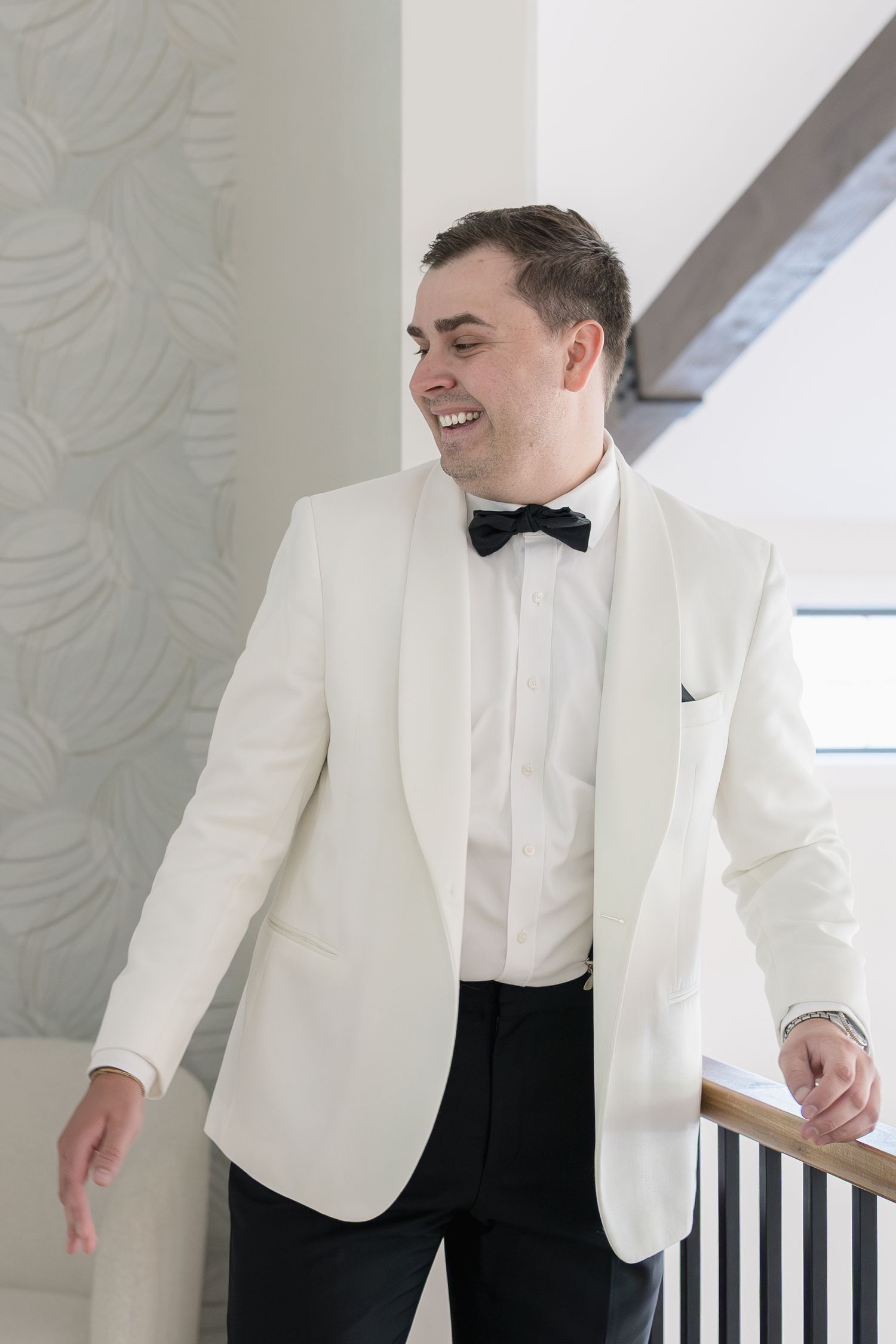 Smiling groom in white tuxedo jacket with black bow tie and pants standing on staircase.