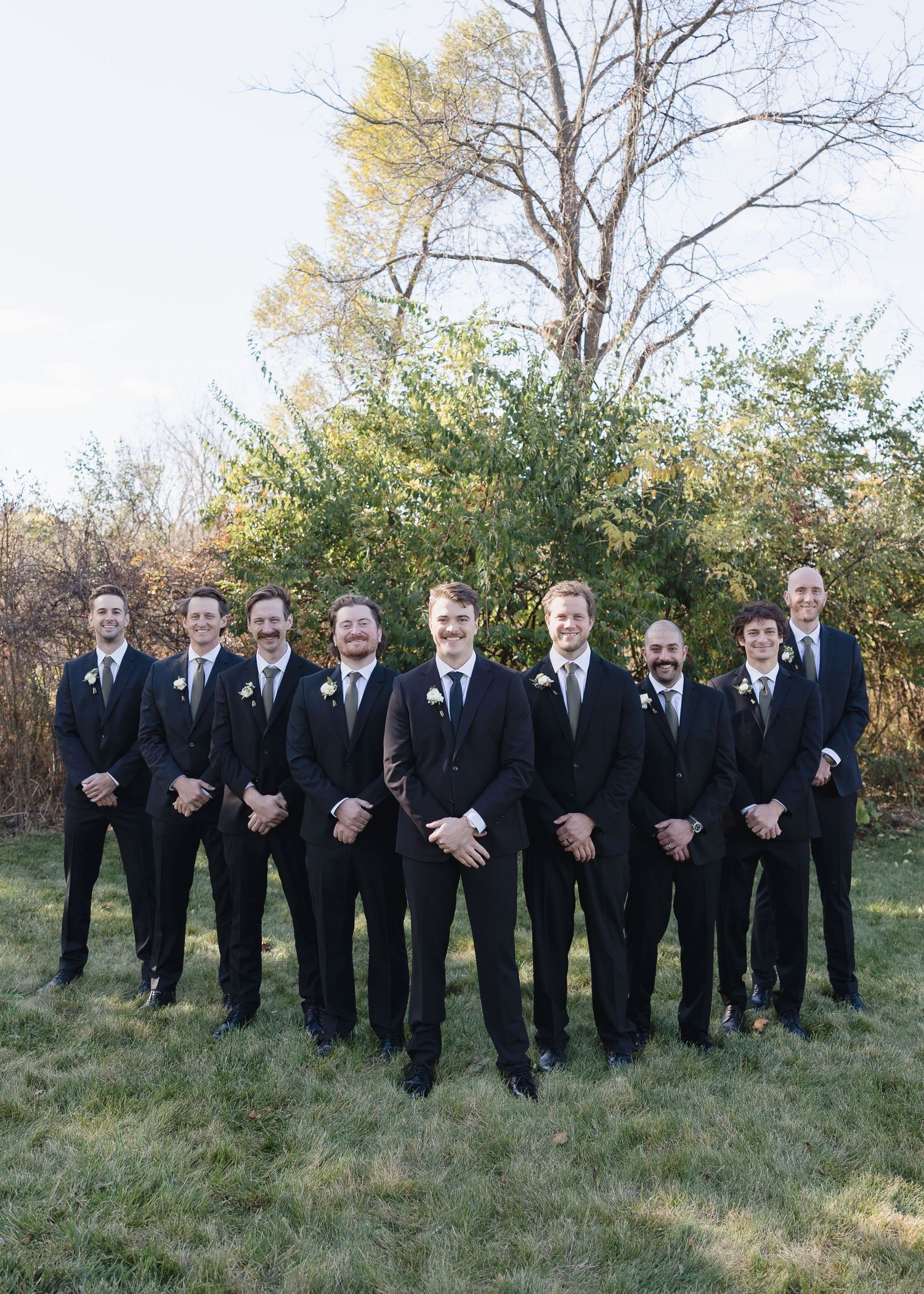 Groomsmen in matching black suits and ties pose formally for group photo outdoors.