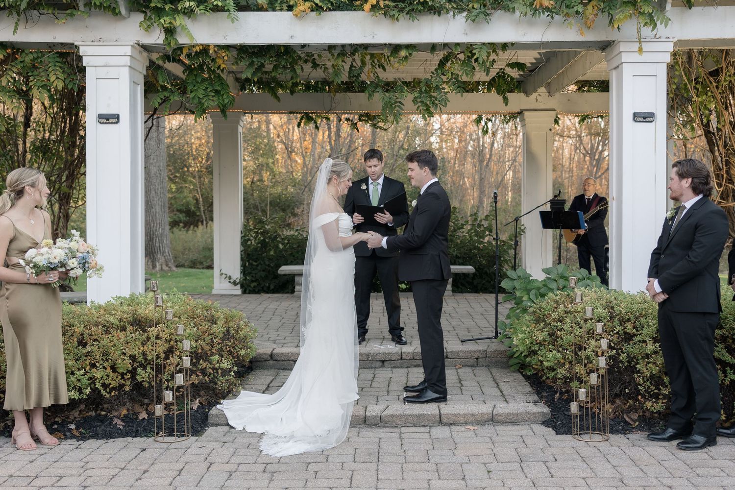 A wedding ceremony under a white pergola decorated with climbing vines.