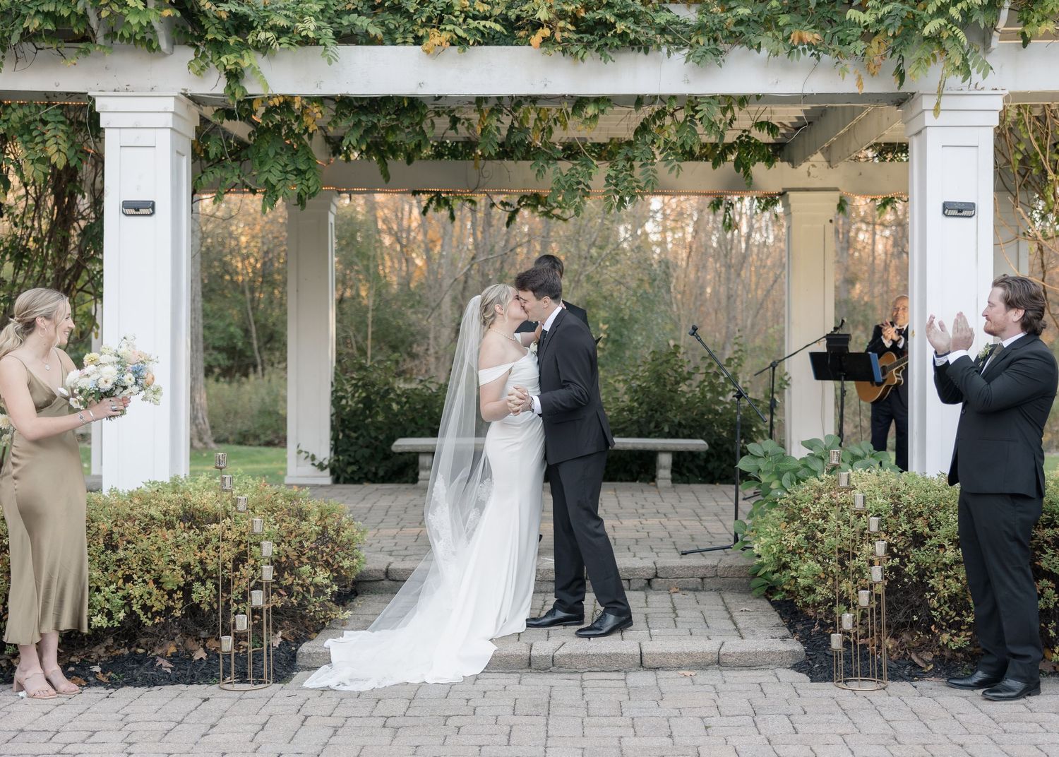 A wedding ceremony taking place under a white pergola decorated with greenery and vines.