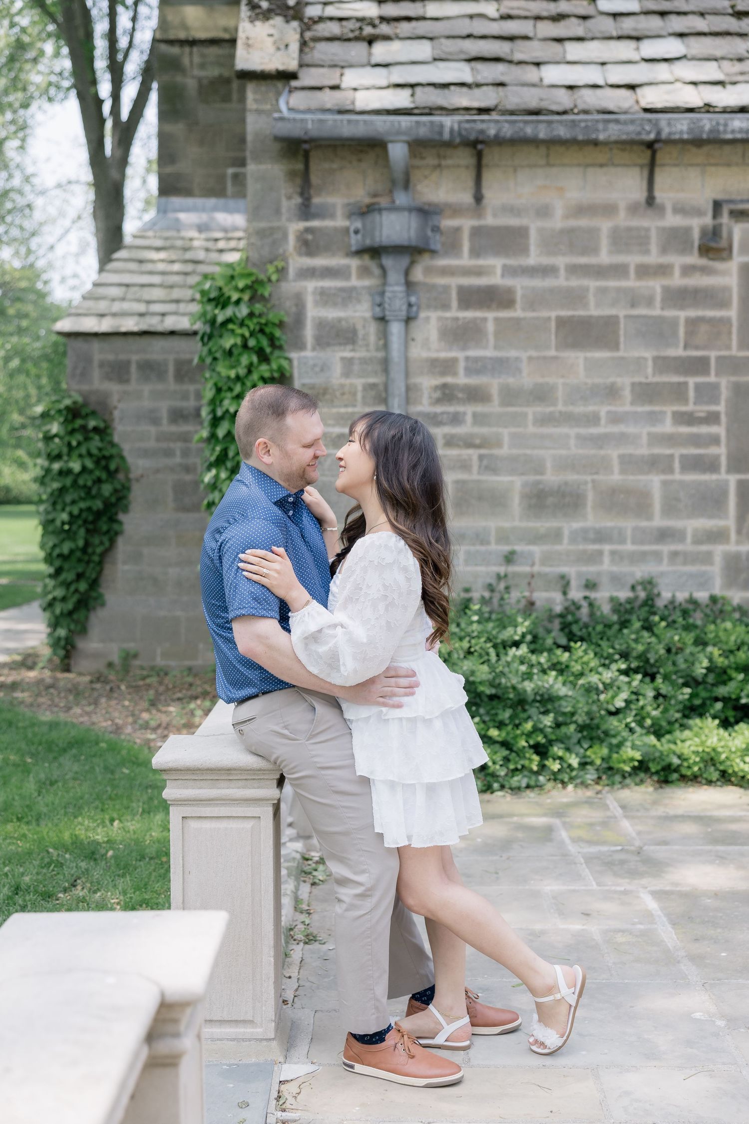 A romantic couple poses together near a stone building with ivy during an engagement photoshoot.