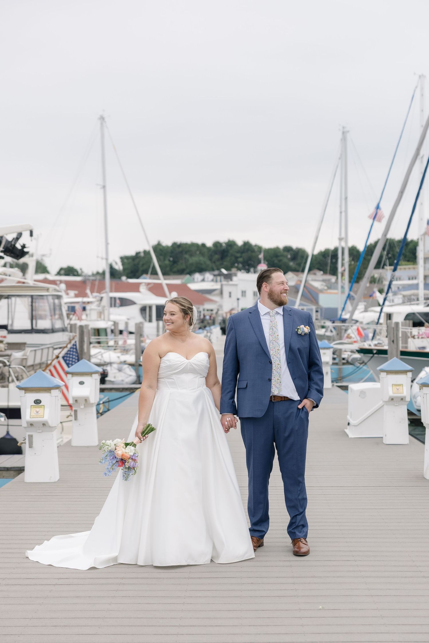Bride and groom walk hand-in-hand on a marina dock, she in a white gown, he in a blue suit.